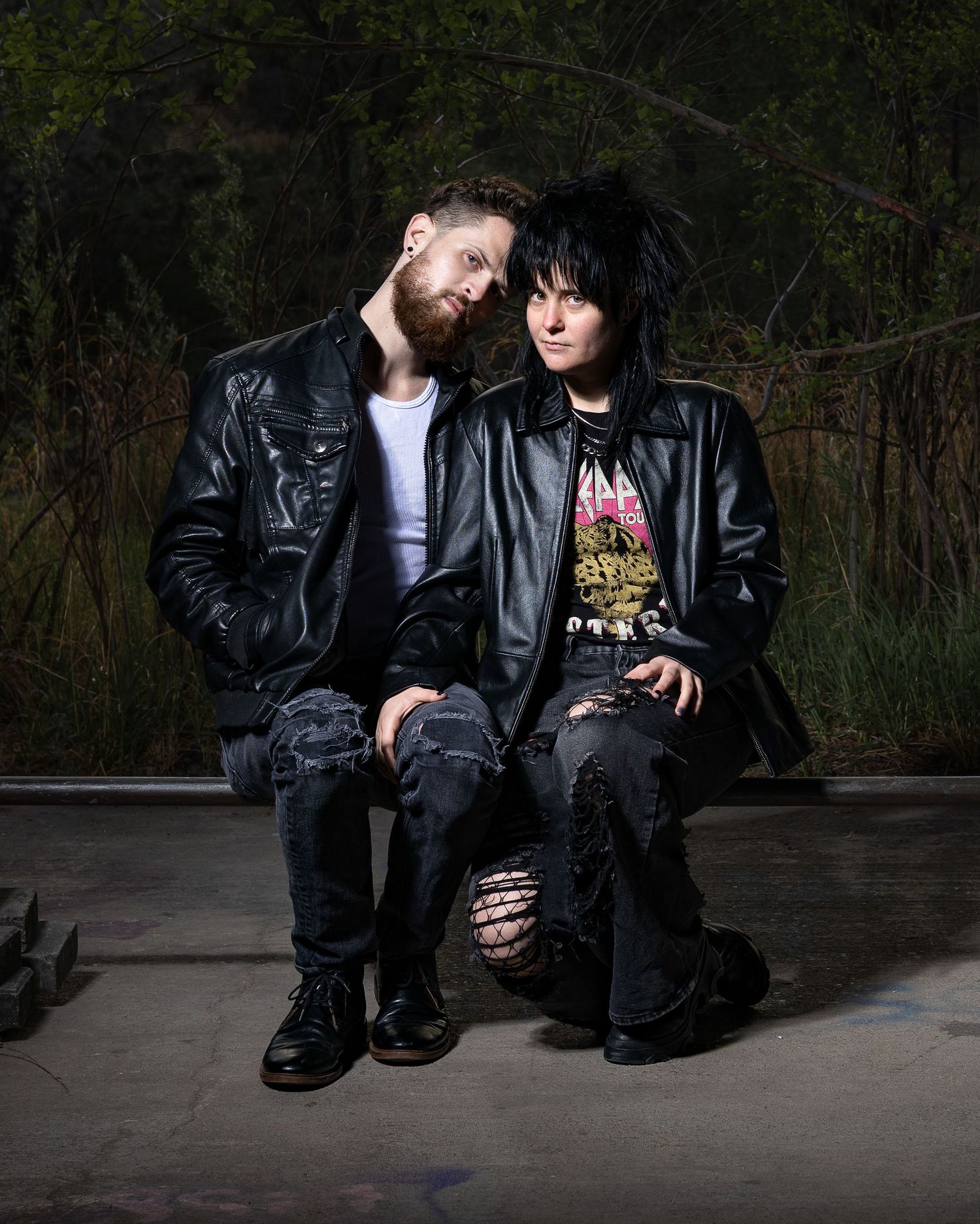 Two people in black leather jackets and edgy fashion sit together outdoors in a moody, alternative-style portrait.