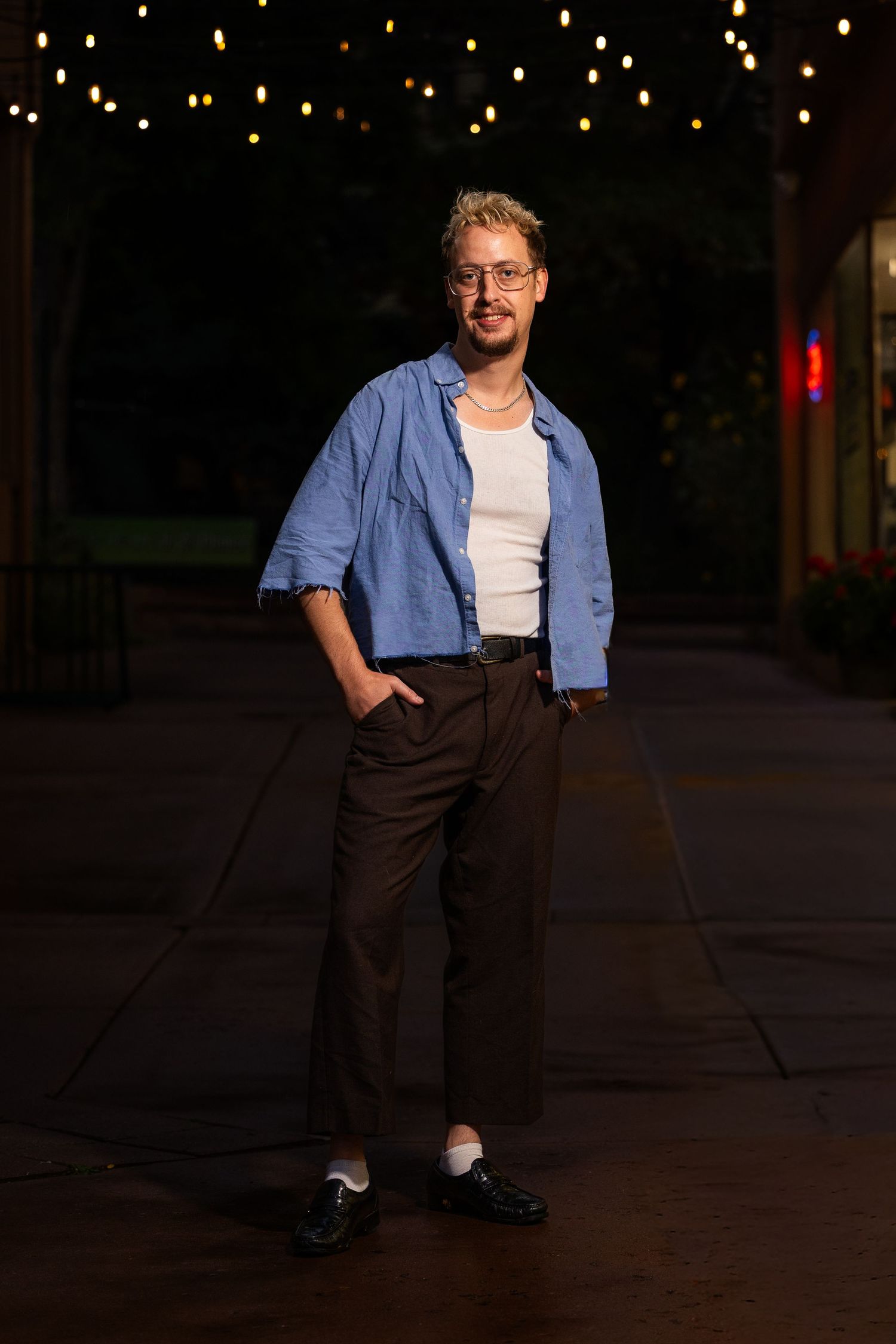 Person in blue shirt and white top poses in a dark outdoor setting with glowing lights in the background.