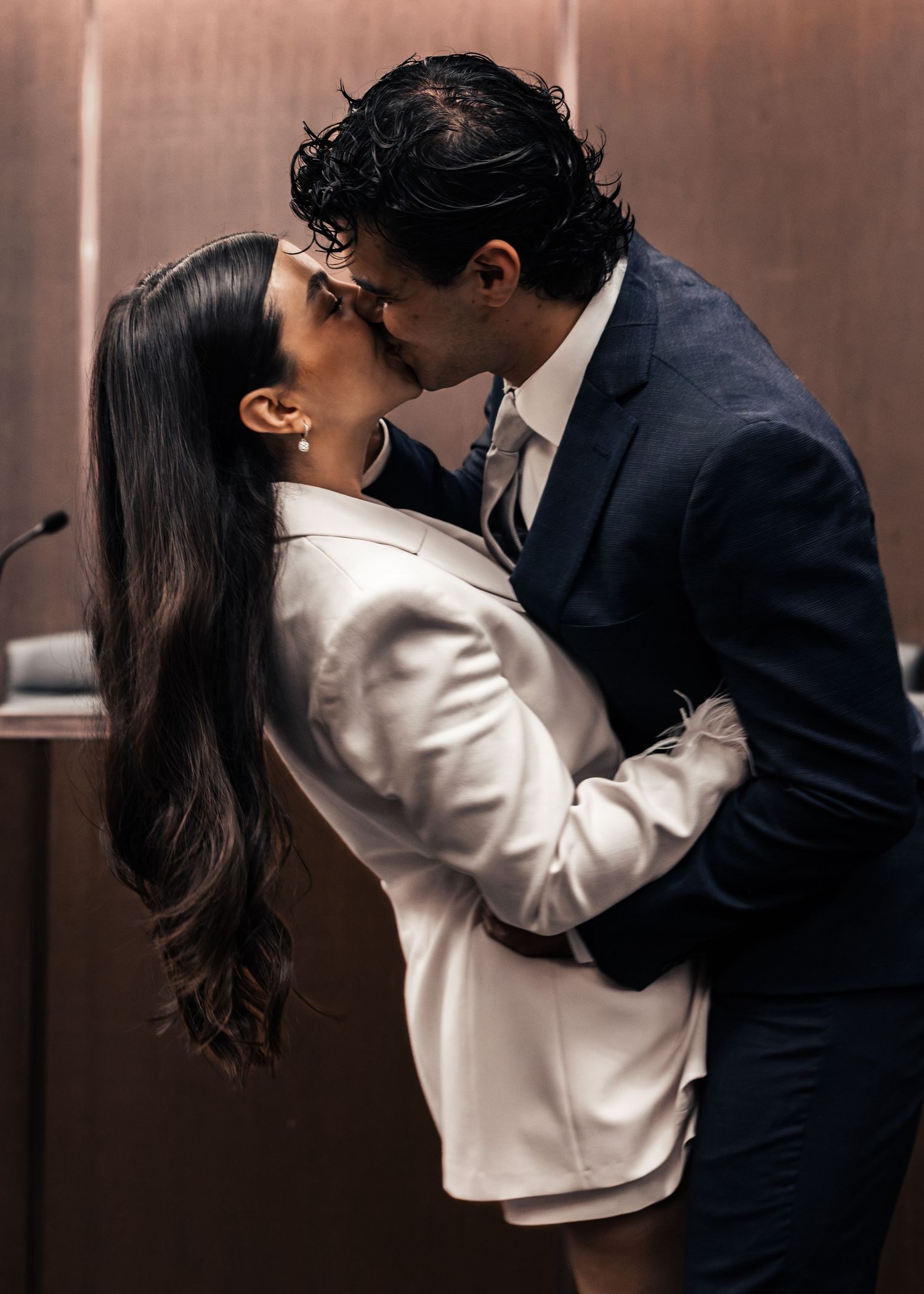 Couple sharing a romantic kiss in a courthouse, dressed in formal attire, white blazer and navy suit.