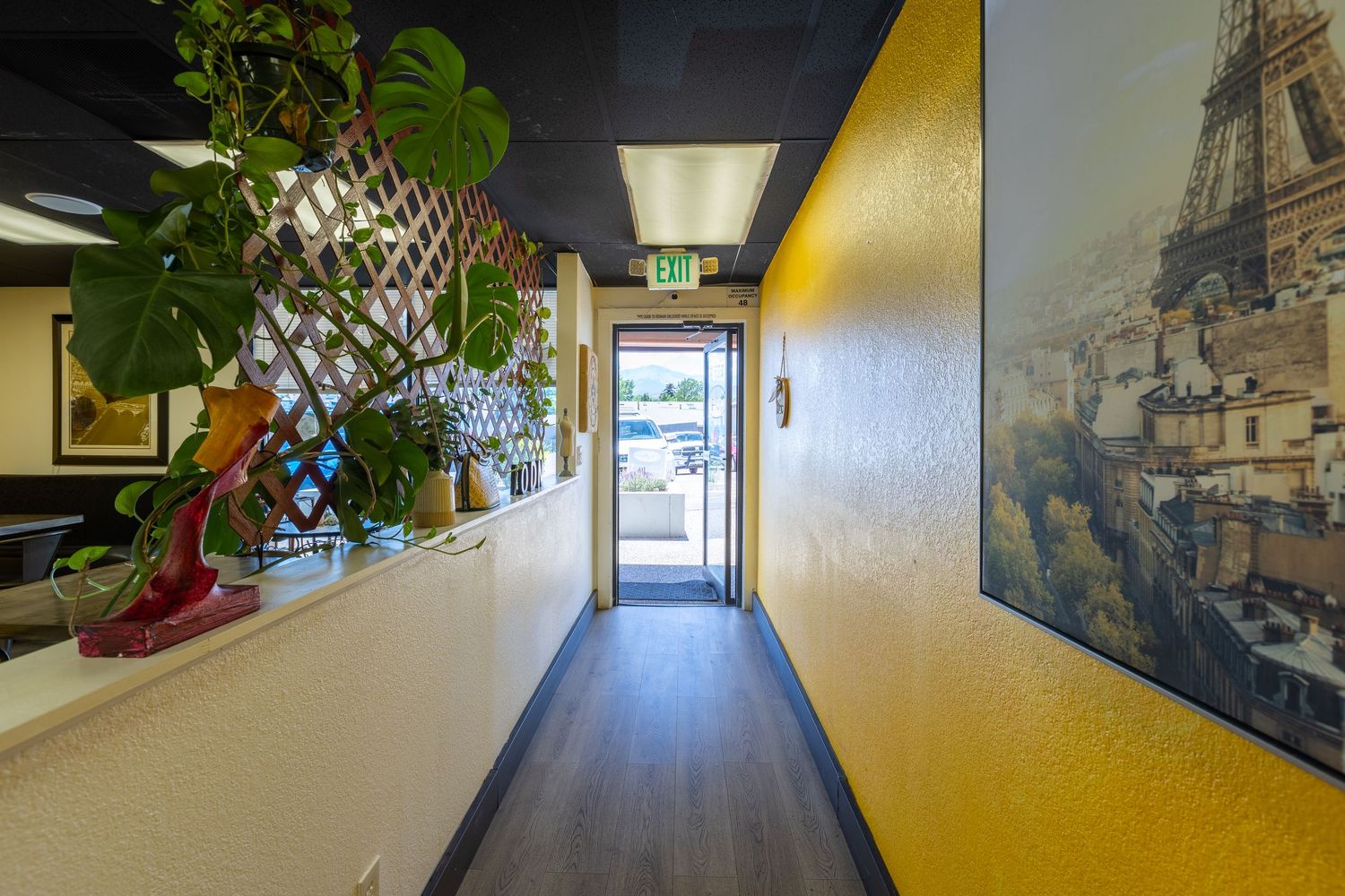 Narrow hallway with yellow walls, Eiffel Tower mural, hardwood floors, and lush green plants leading to a glass door.