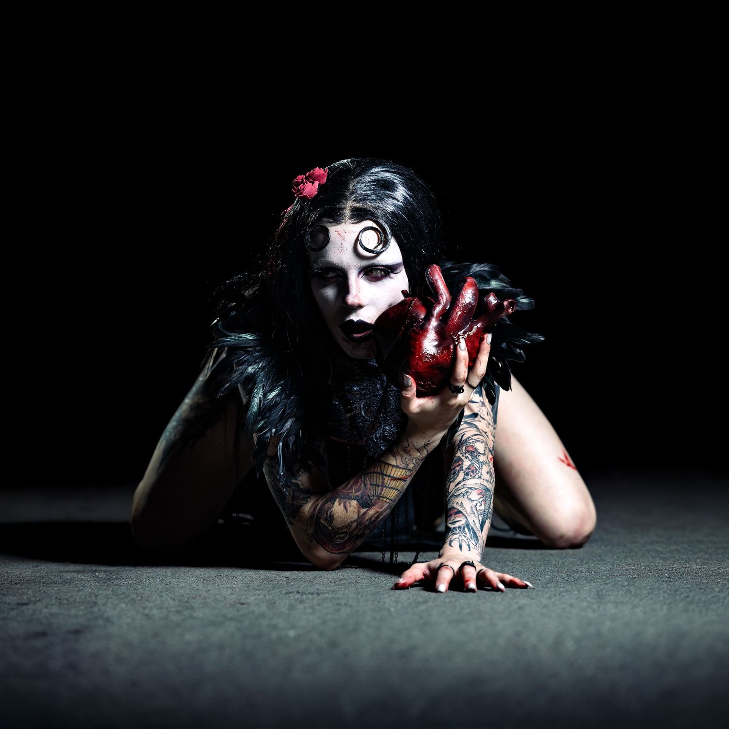A tattooed performer in dark gothic makeup with black feathers crawls dramatically on the floor with blood-red hands.