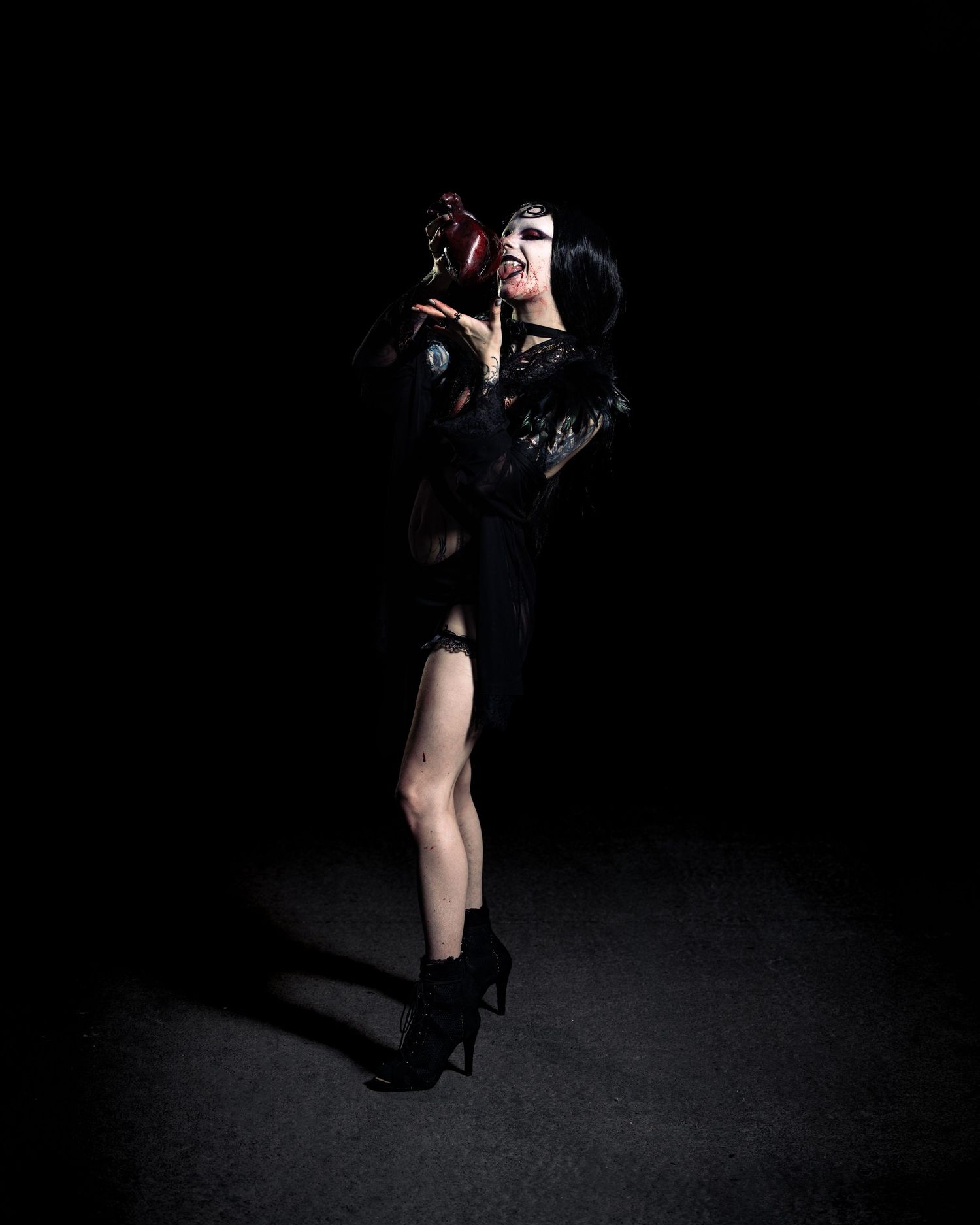 Gothic performer in black outfit and heels holds a dark floral prop against a dramatic black background.