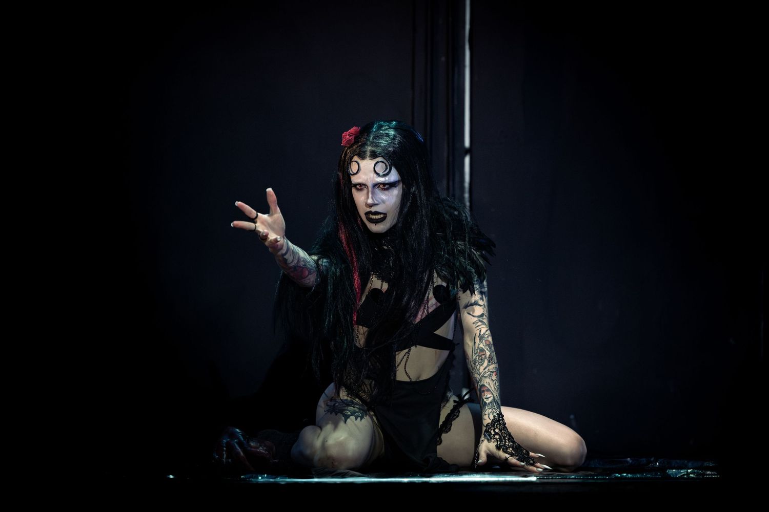 Gothic performer with elaborate face paint and tattoos kneels dramatically on a dark stage.