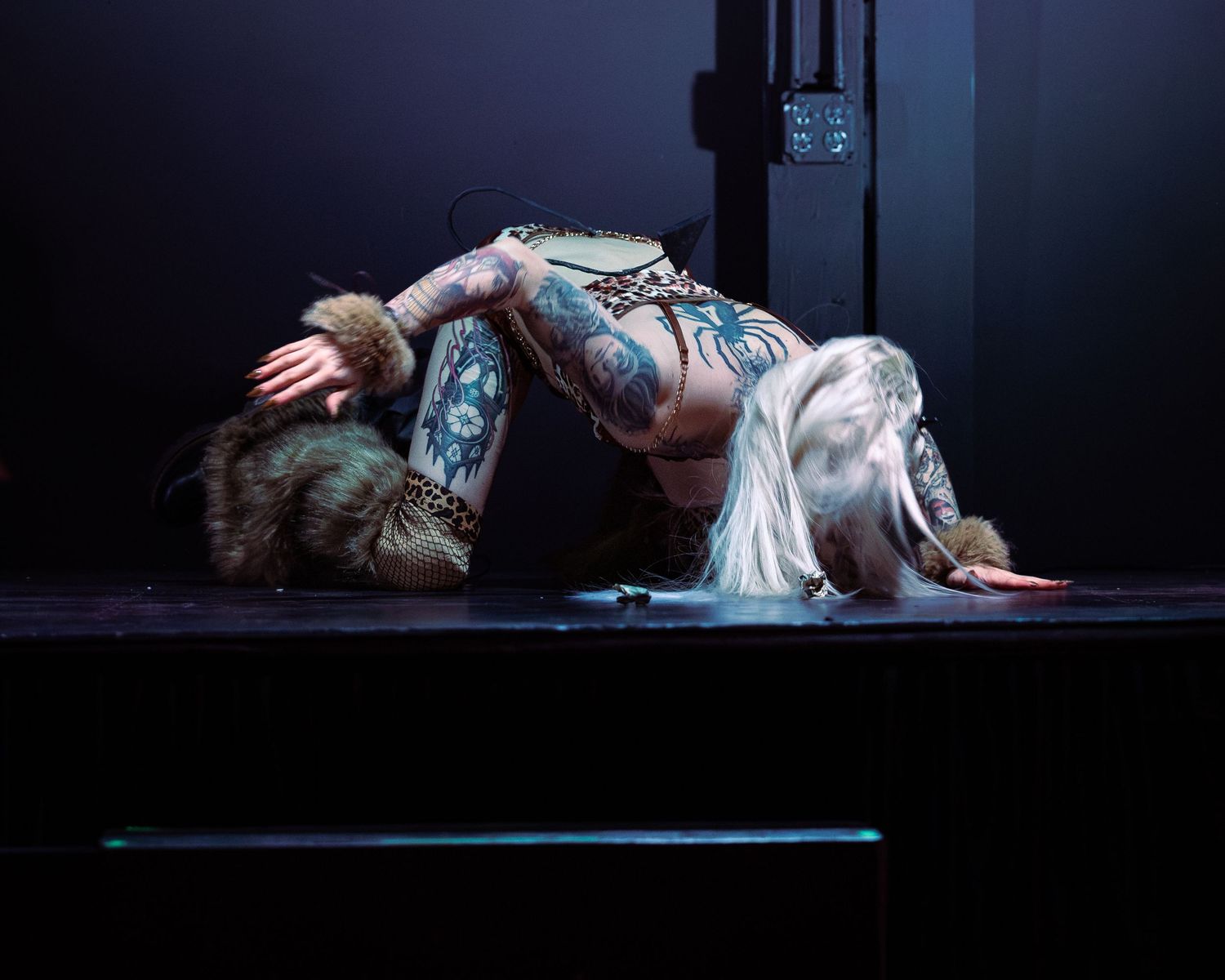 Heavily tattooed performer bending dramatically on a dark stage during an artistic performance.