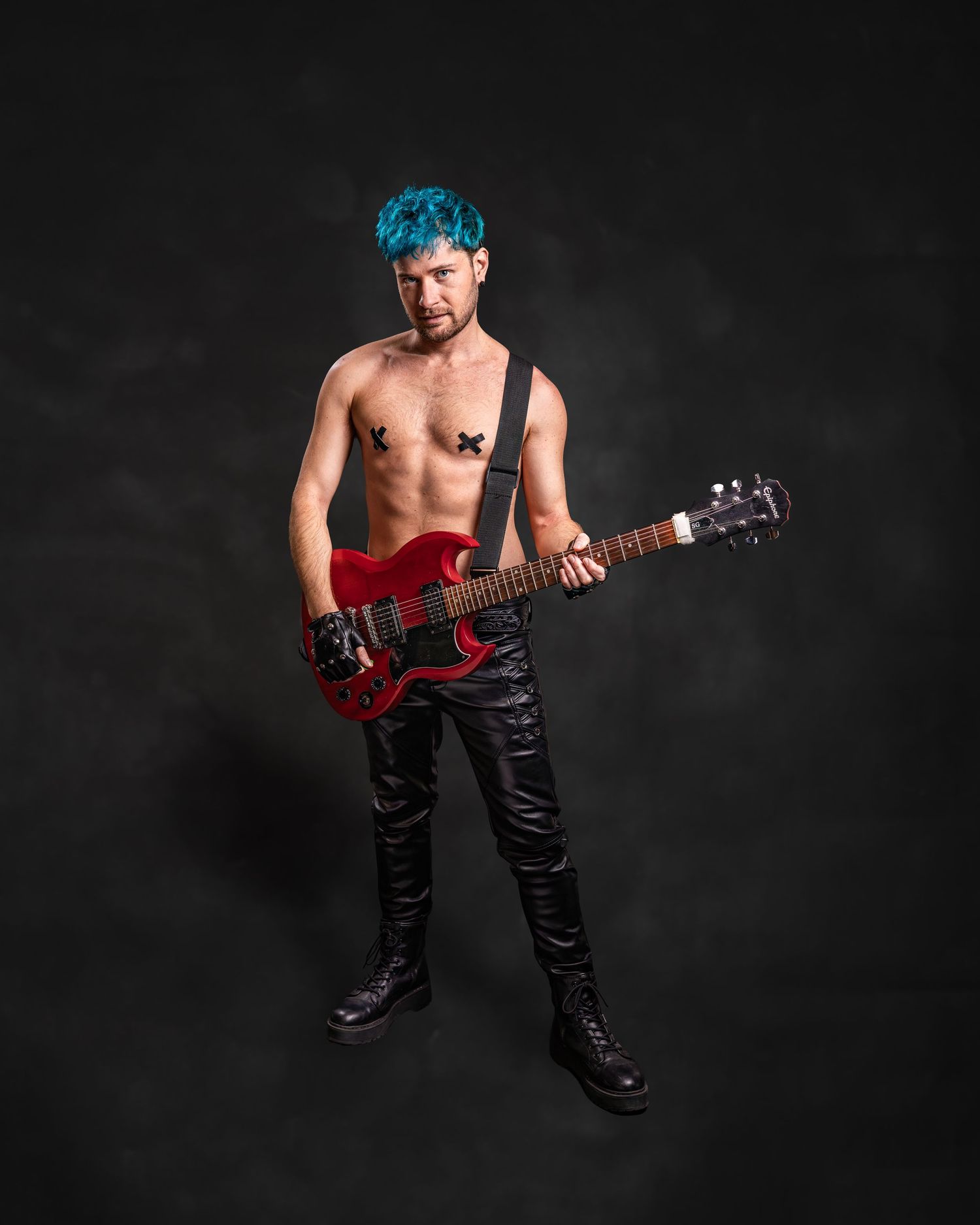 Shirtless musician with teal hair playing red electric guitar, wearing black leather pants and boots on dark background.