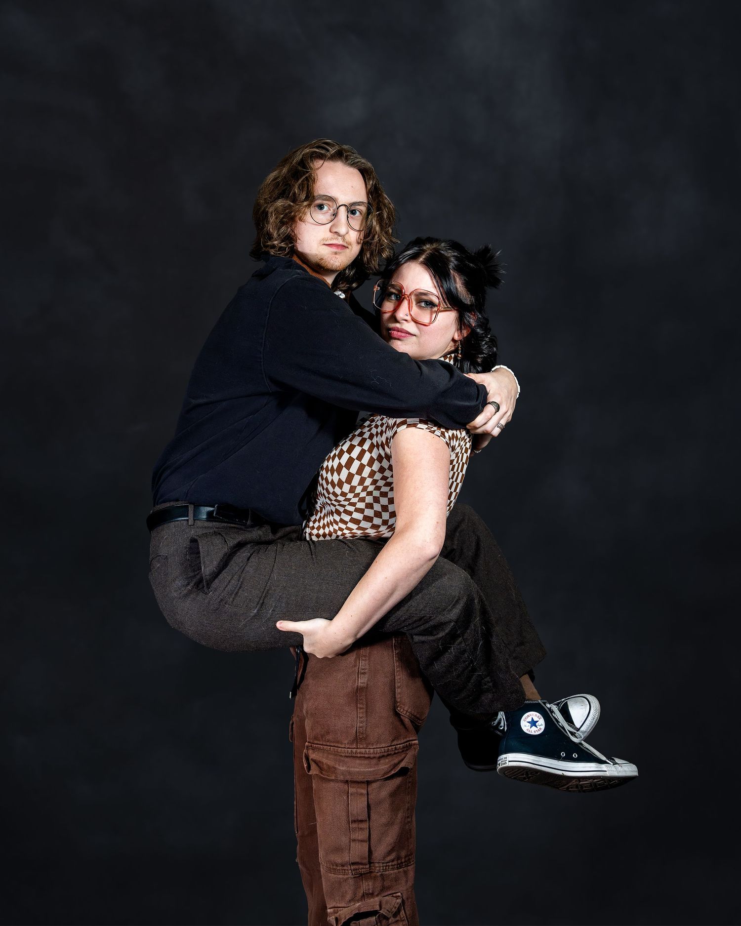 Two young people posing for a studio portrait, one giving the other a piggyback ride, both wearing glasses.