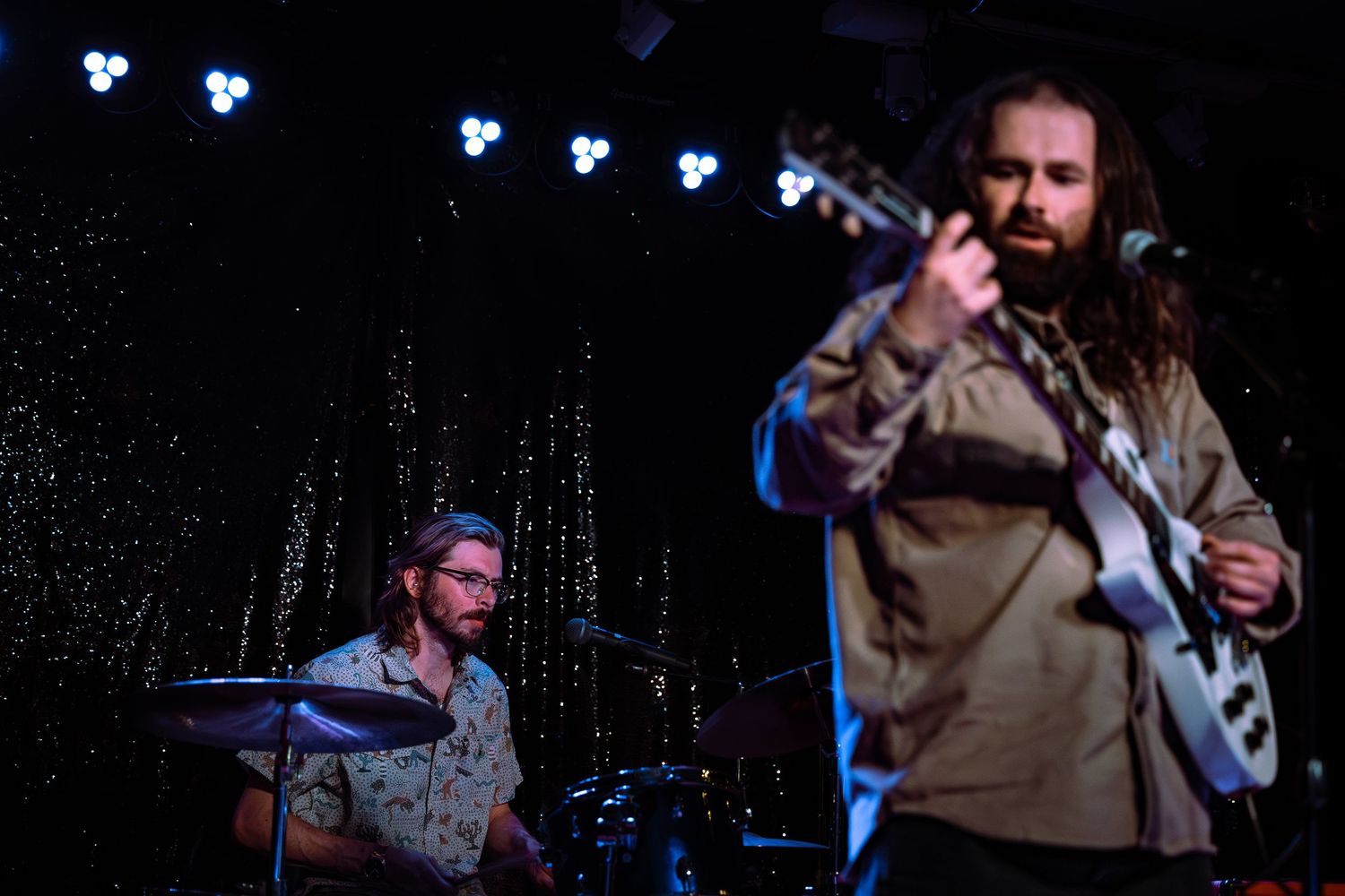 Two musicians performing live, one on guitar and one on drums on stage.