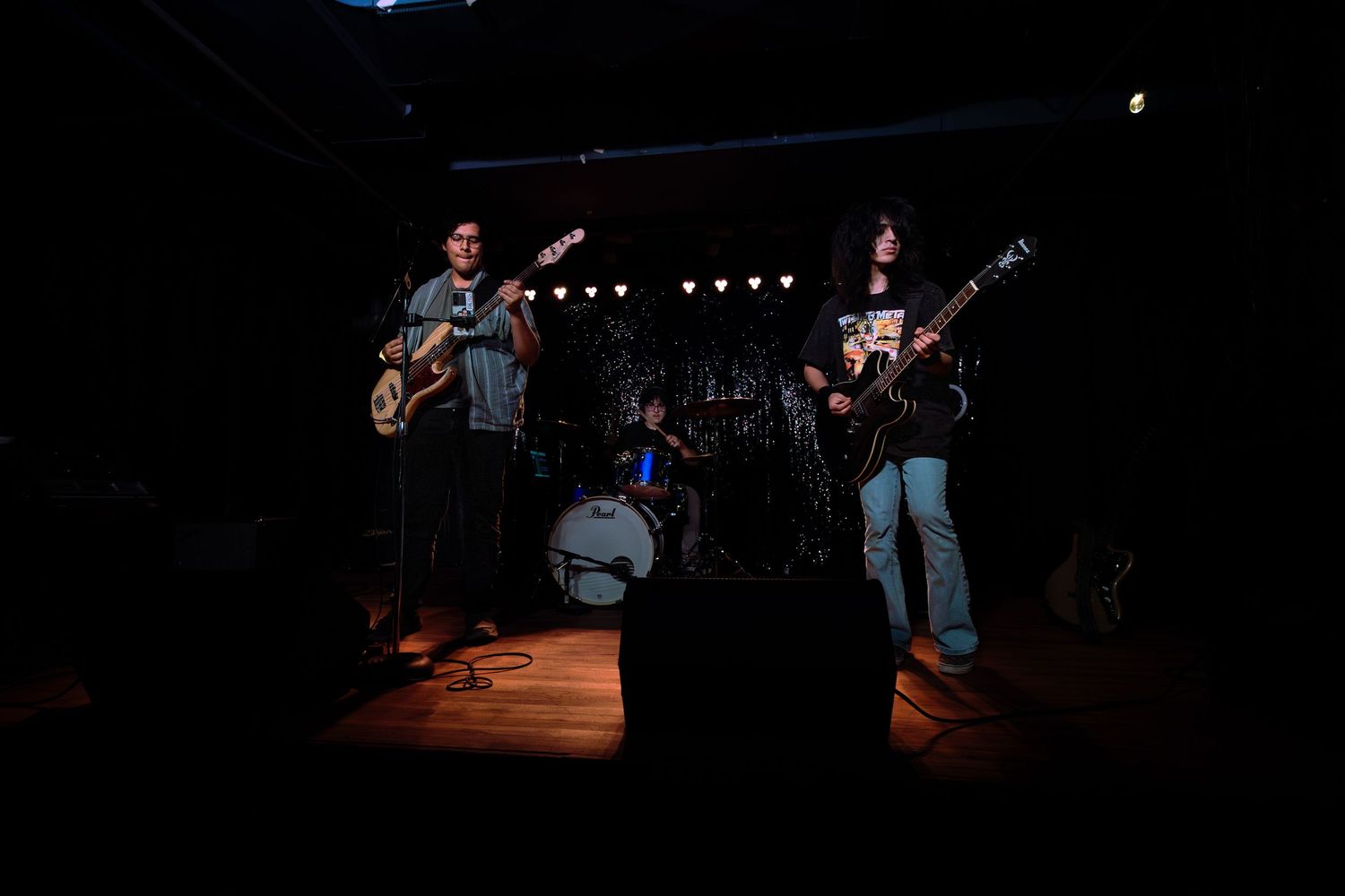 Two guitarists perform on a dimly lit stage with a drummer in the background at a live music venue.