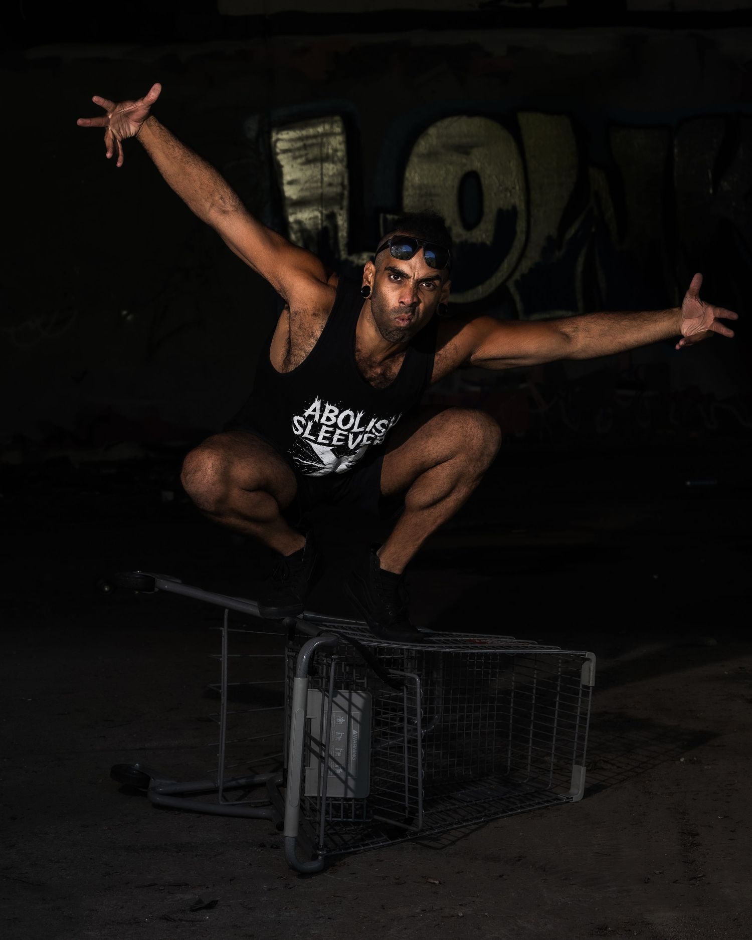 Athletic performer in black tank top executes dramatic jump pose over metal cart in dark urban setting.