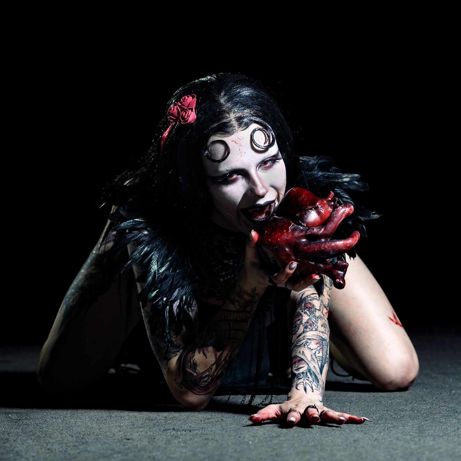 Gothic woman with dark makeup, tattoos, and dramatic costume poses on floor holding red prop in dark studio setting.