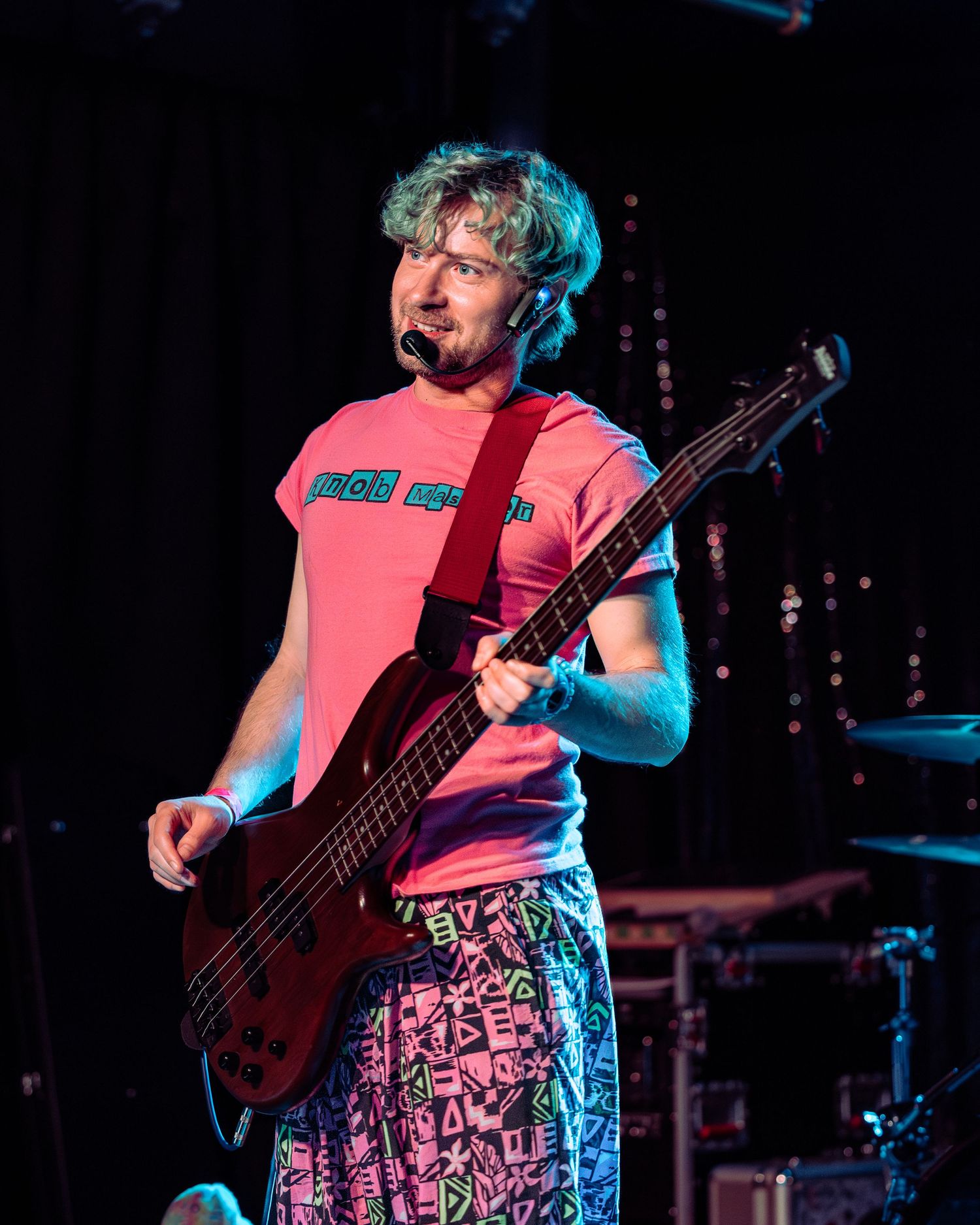 Male bassist in pink tee and patterned shorts performs on stage with red bass guitar.