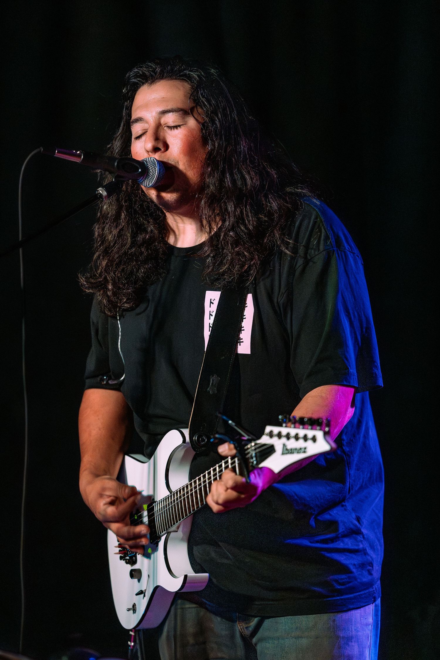 Long-haired male guitarist singing into microphone on stage holding a white electric guitar.