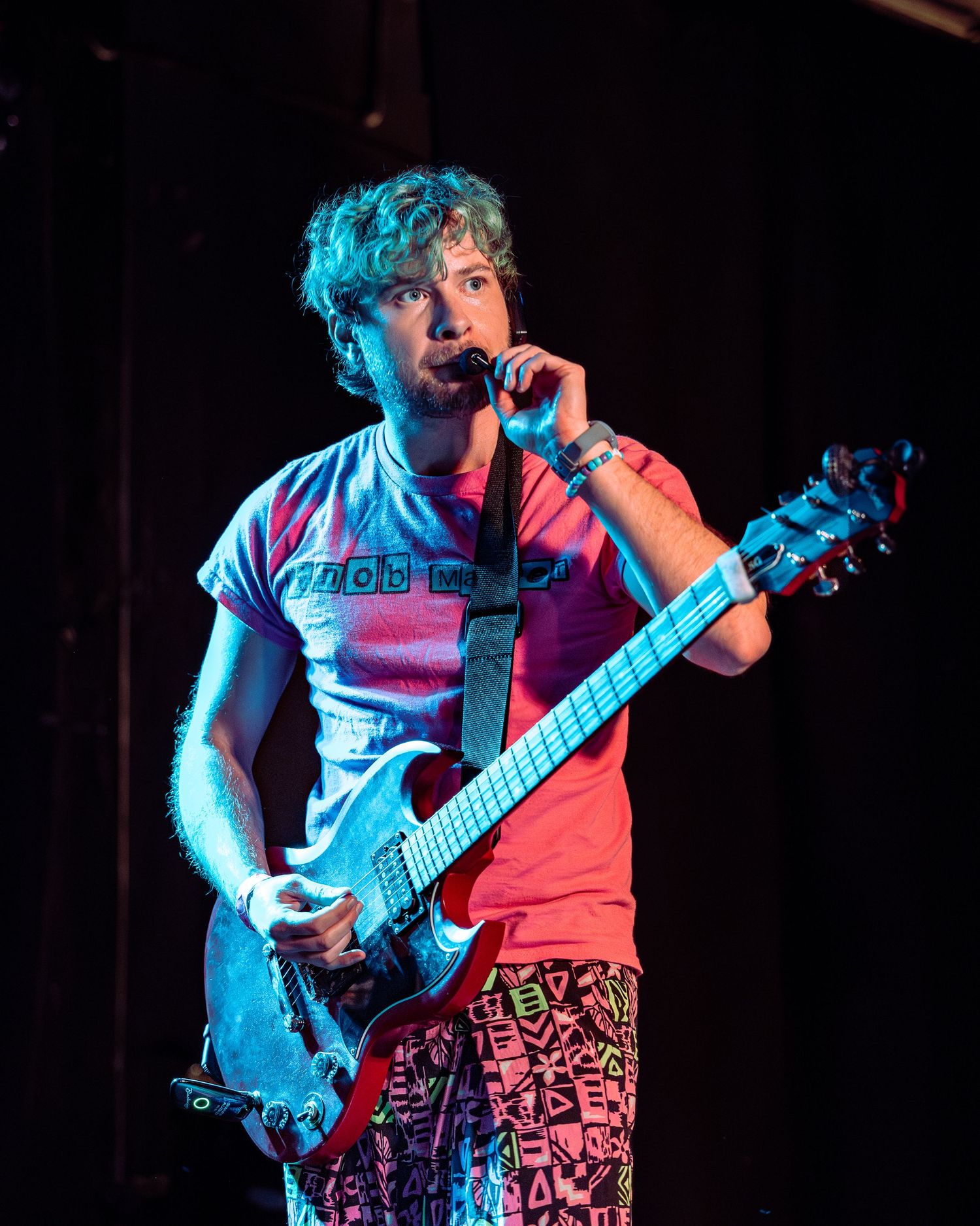 Male guitarist in pink shirt and colorful pants sings into microphone while performing live on stage.