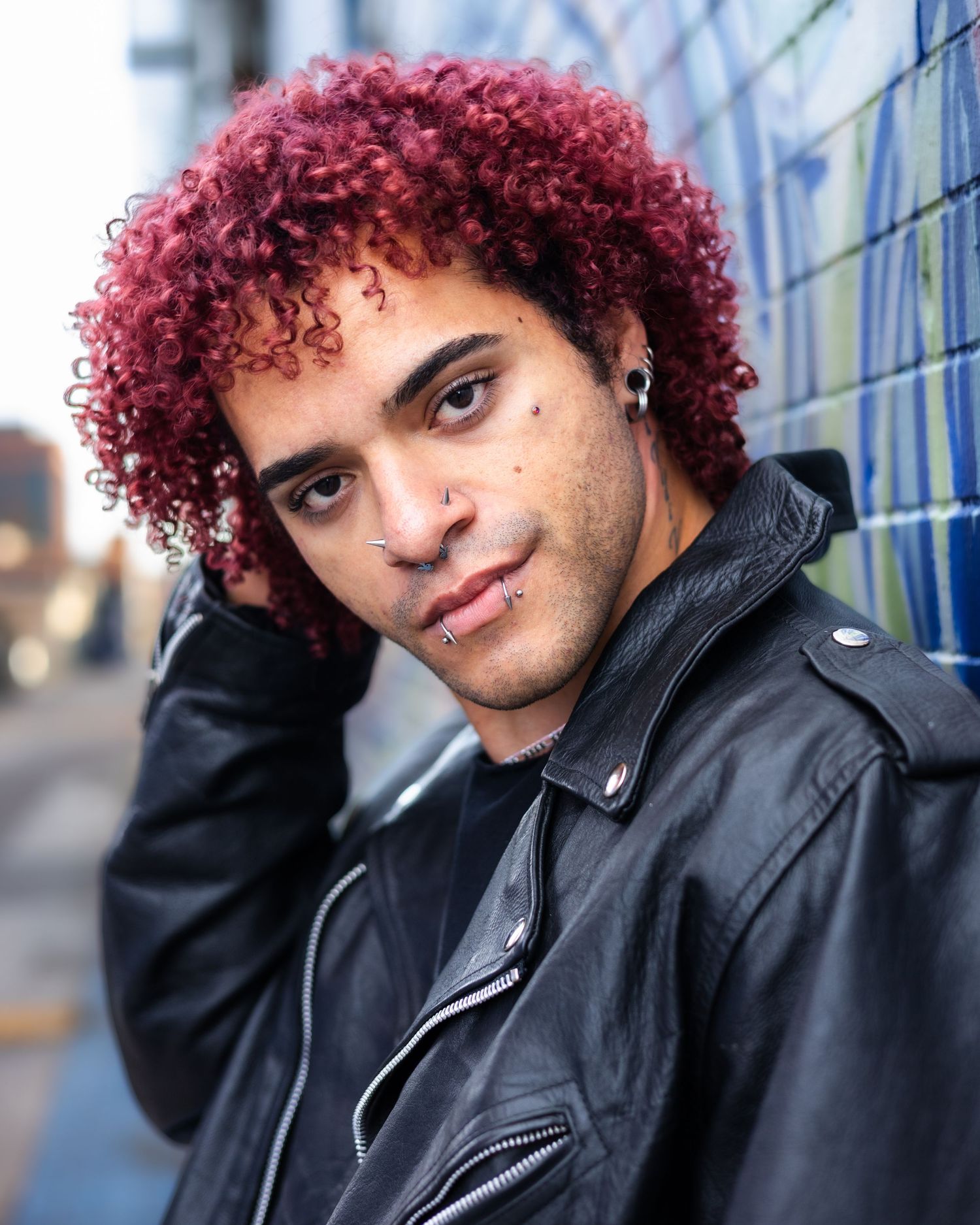 Person with vibrant red curly hair wearing a black leather jacket poses against an urban backdrop.