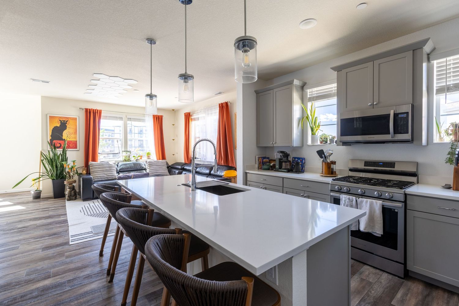 Modern kitchen with gray cabinets, white island, pendant lights, stainless appliances, and open living area with orange curtains.