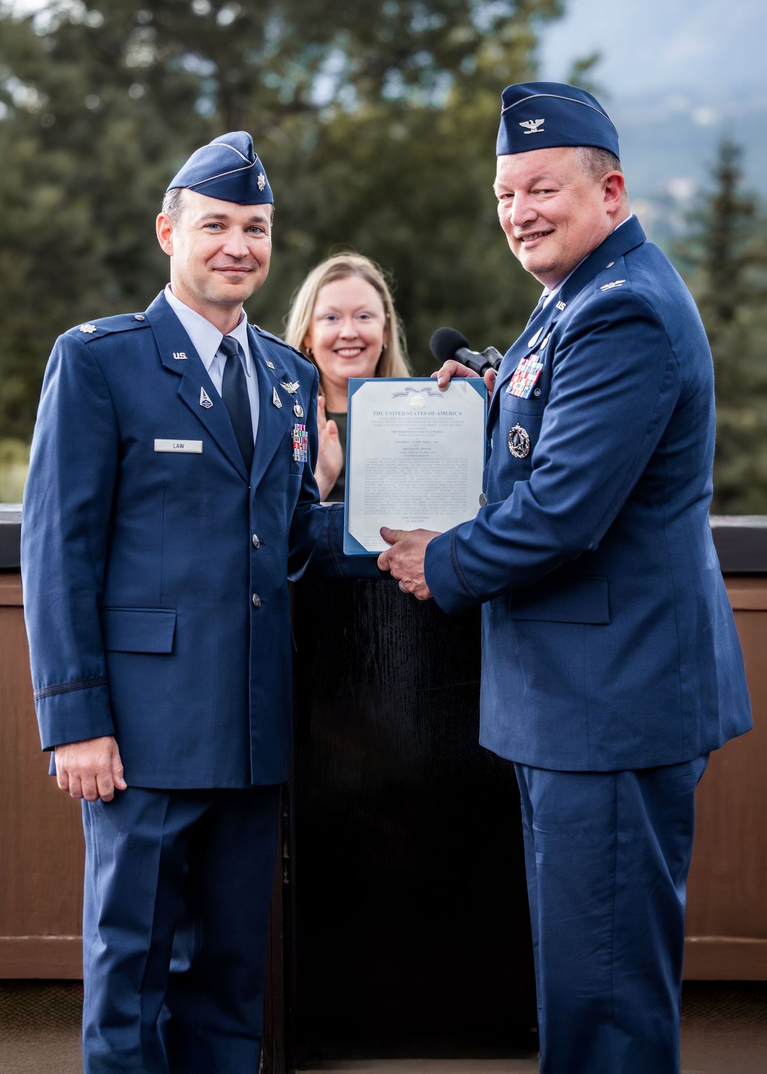 Air Force service members in dress blues present an official document during a formal ceremony outdoors.