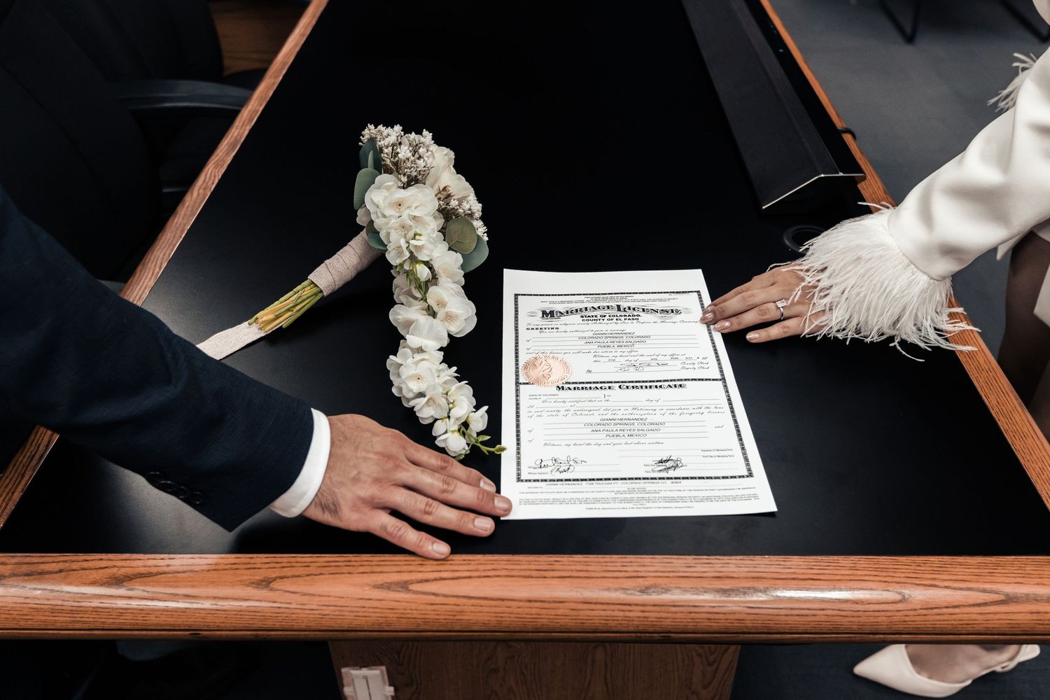 Couple signing a marriage certificate on a dark table with a white floral bouquet beside it.