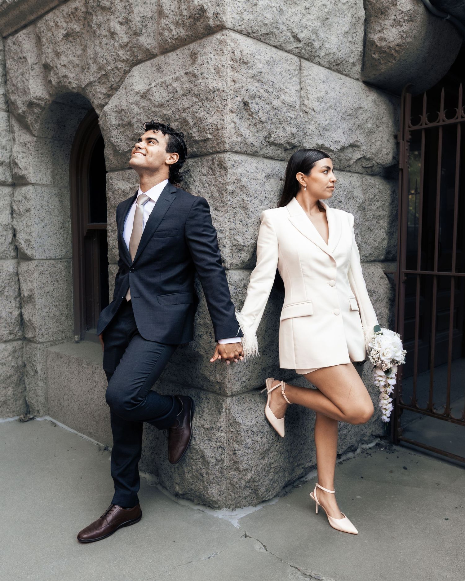 Stylish couple holding hands in wedding attire against stone wall, woman in white blazer dress, man in dark suit.