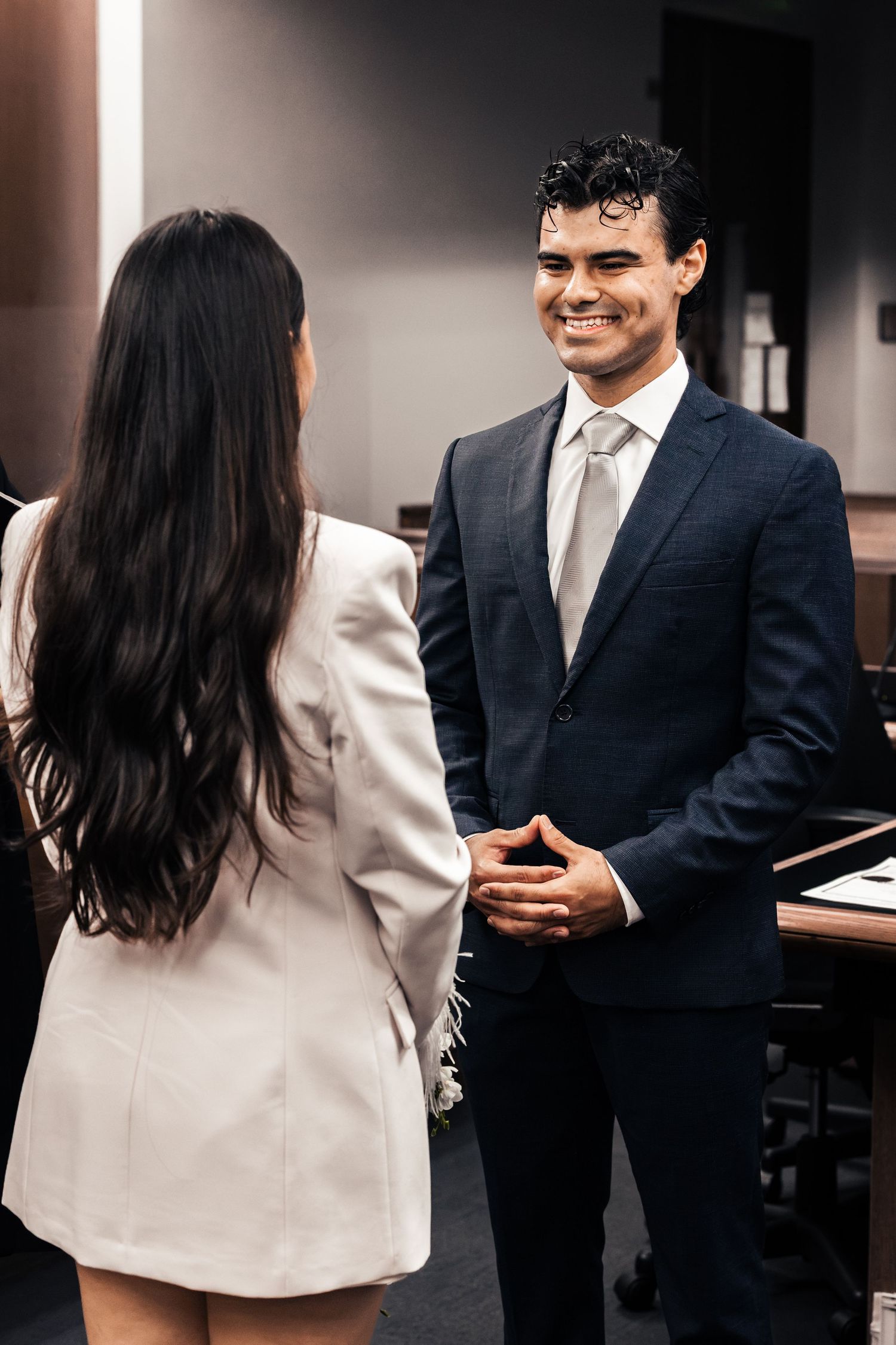 A smiling man in a navy suit and tie converses with a woman in a white blazer dress indoors.