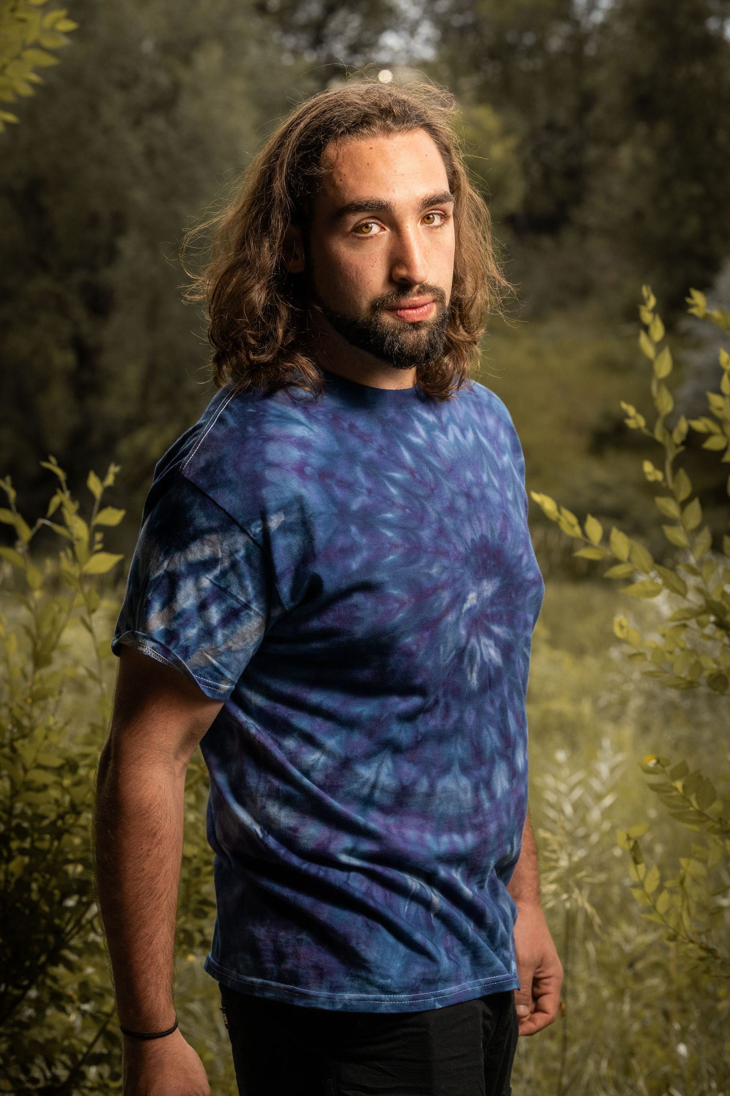 Person wearing blue tie-dye shirt poses outdoors against natural foliage background.