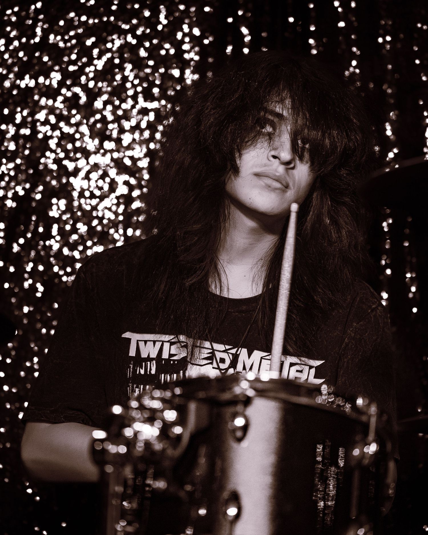 Long-haired drummer playing drums wearing a Twisted Metal t-shirt in a sepia-toned concert photo.