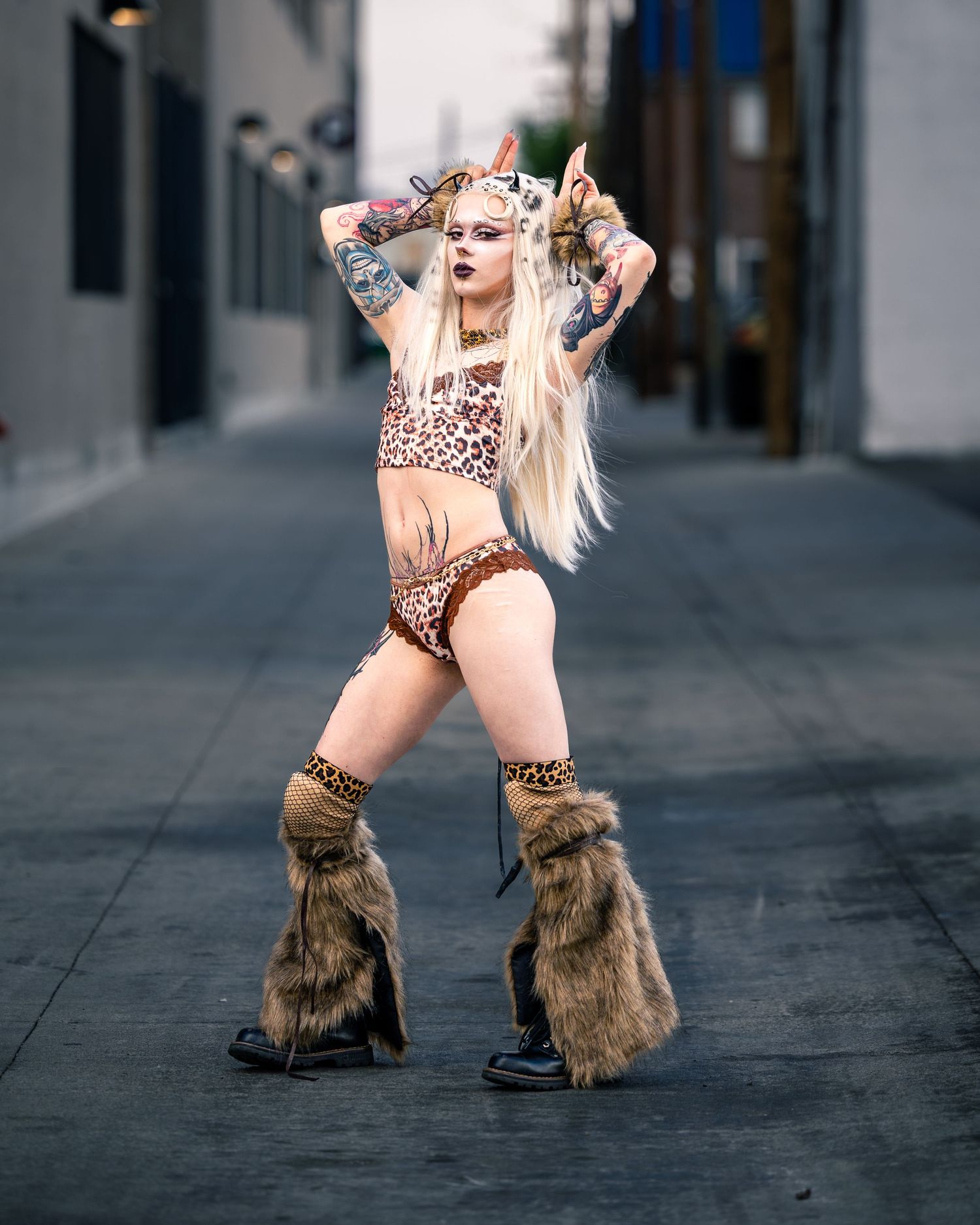 Blonde woman in leopard print costume with horns, fur leg warmers, and heels posing in an urban alley.
