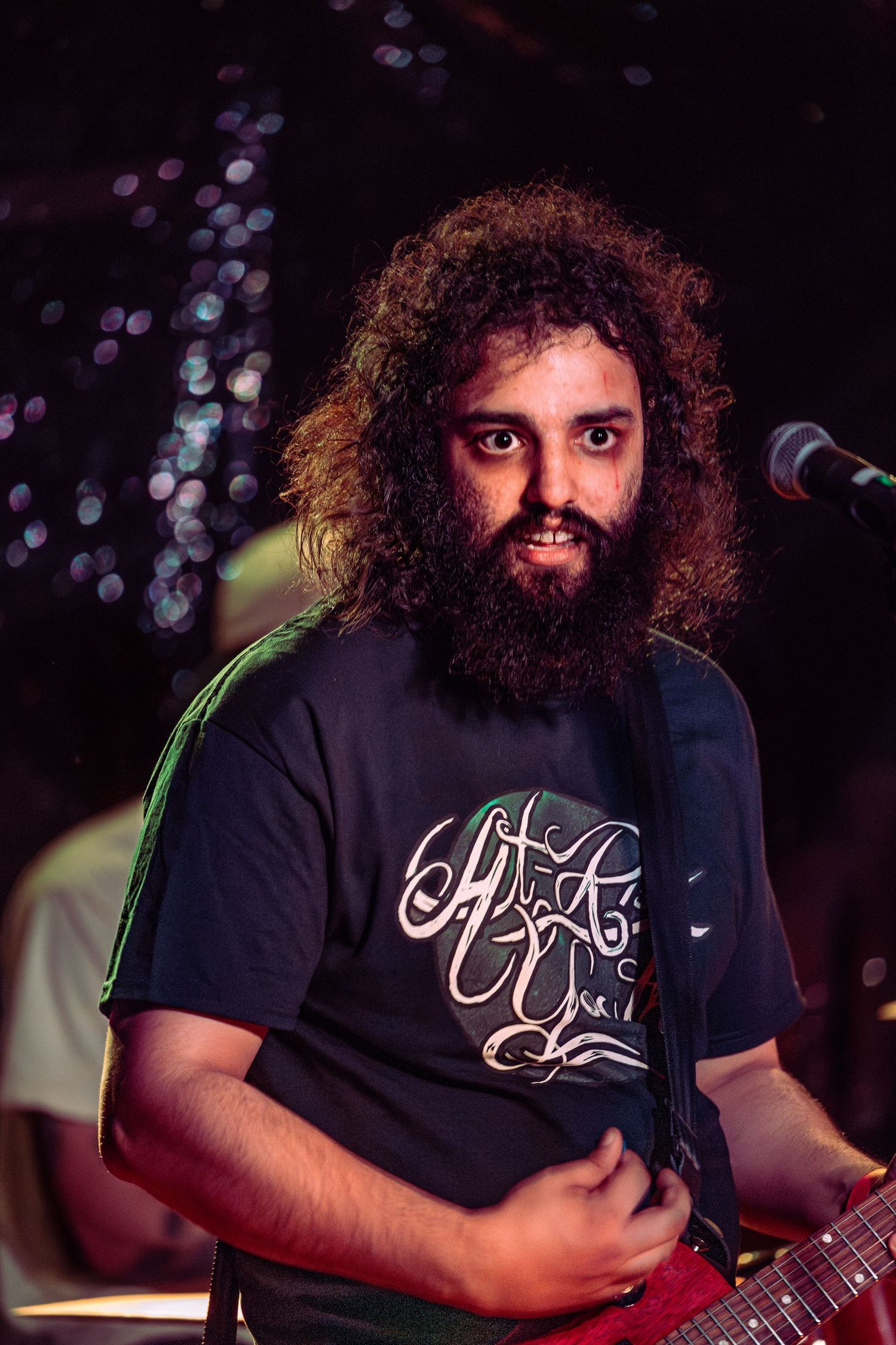 Bearded guitarist with long curly hair smiles on stage wearing black graphic t-shirt.