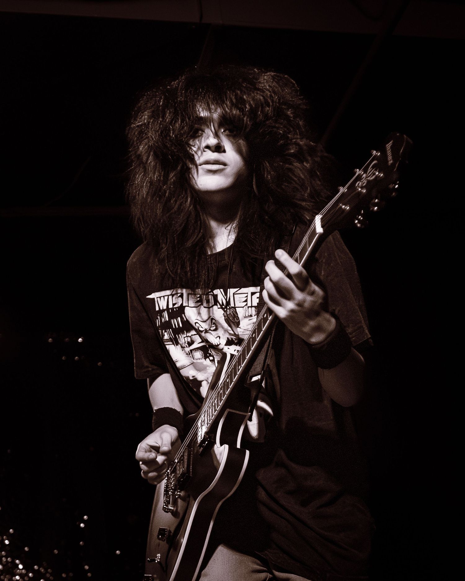 Young male guitarist with long curly hair plays electric guitar on stage in dramatic black and white photo.
