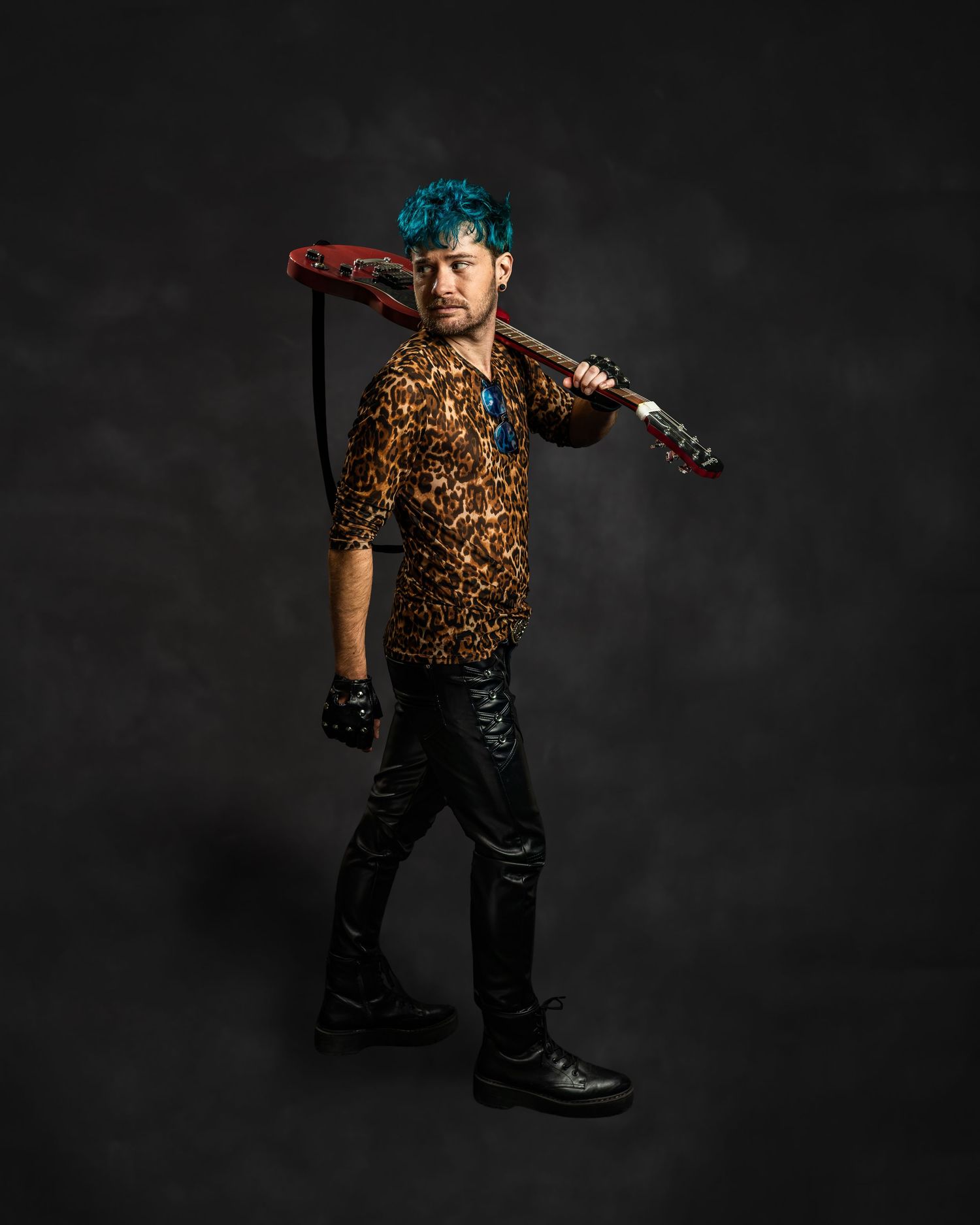 Rock musician with blue hair in leopard shirt posing with red guitar over shoulder on dark backdrop.