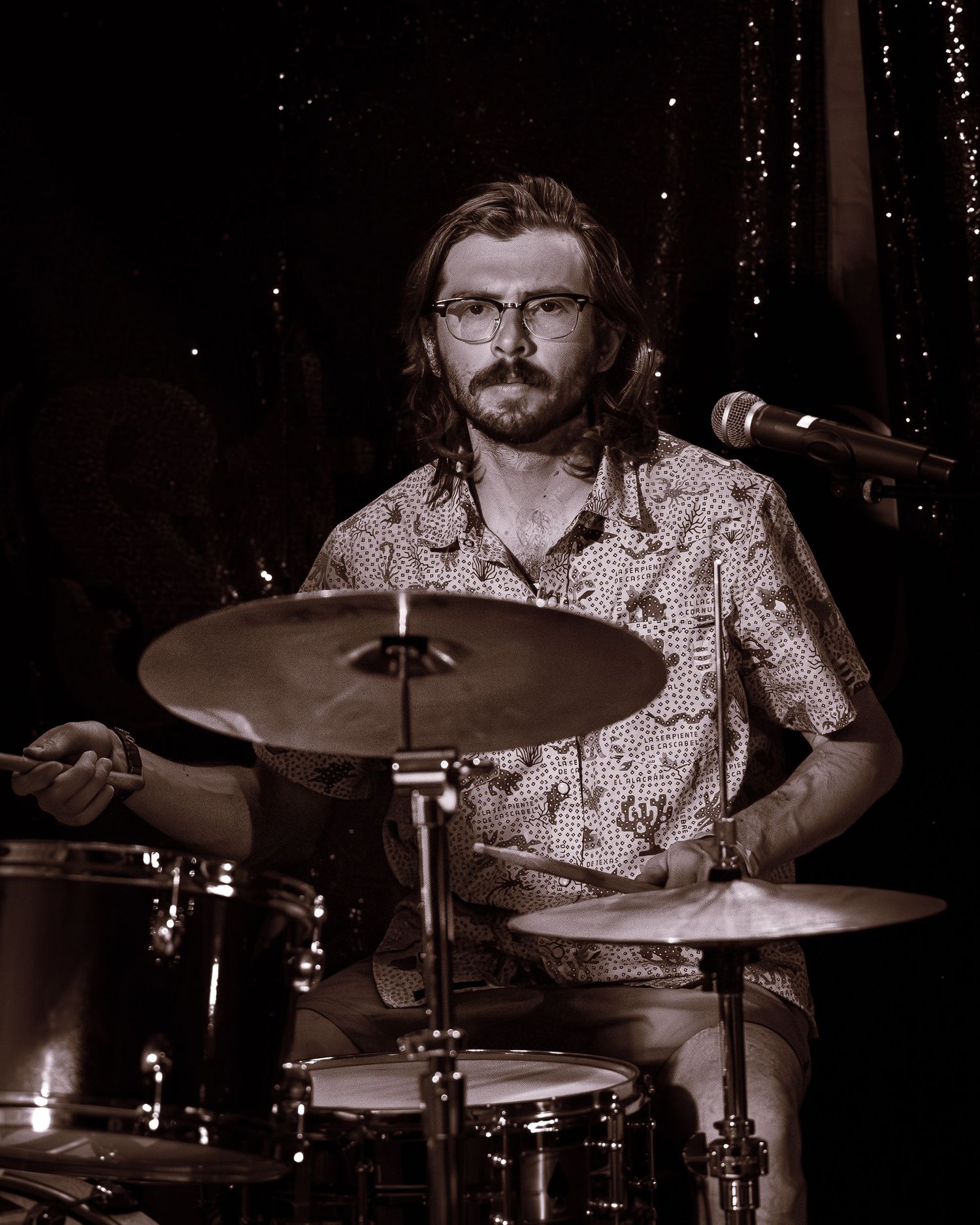 Bearded drummer with glasses playing cymbals in sepia-toned concert photo.