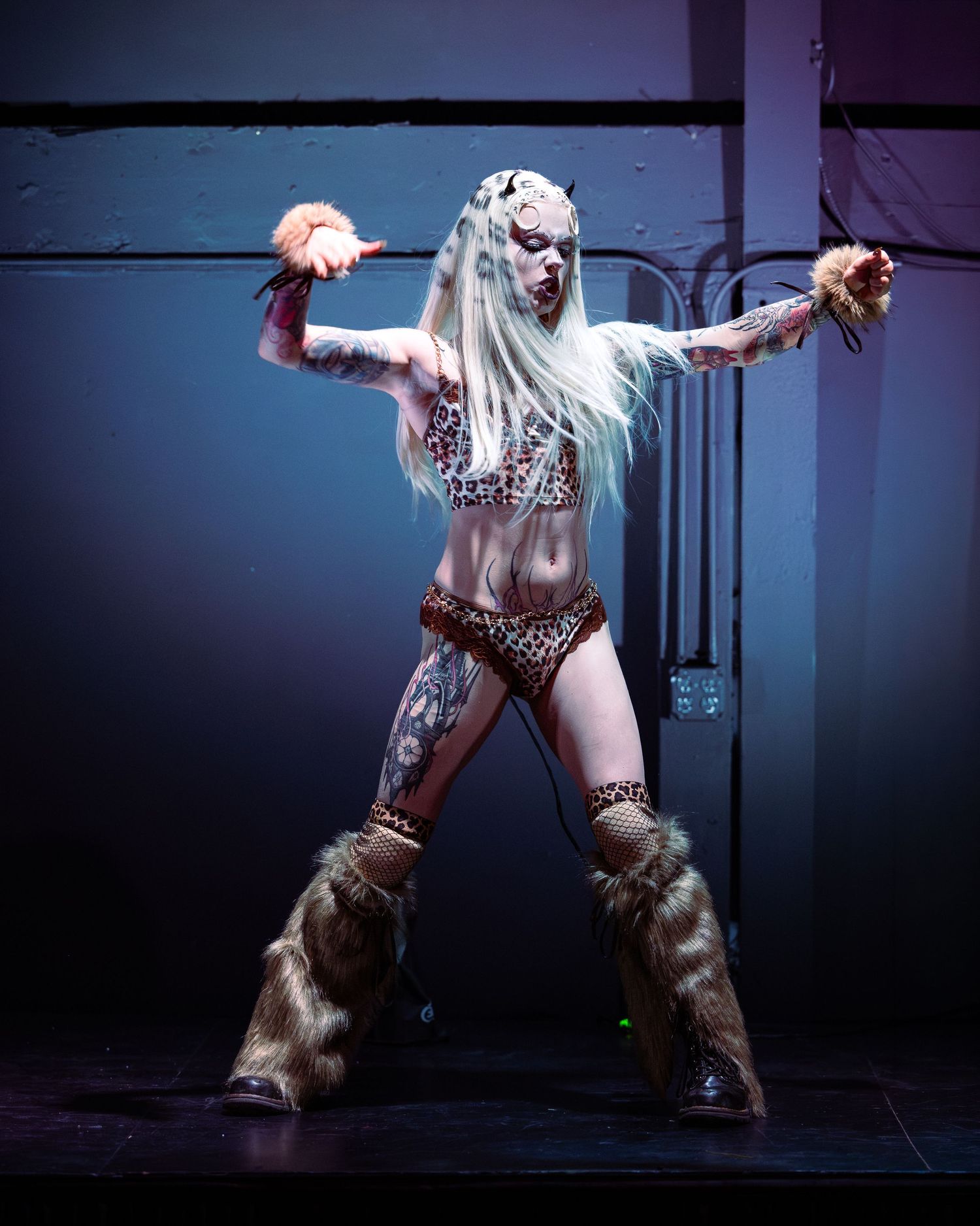 Tattooed performer in leopard print costume with fur boots holds props on stage.
