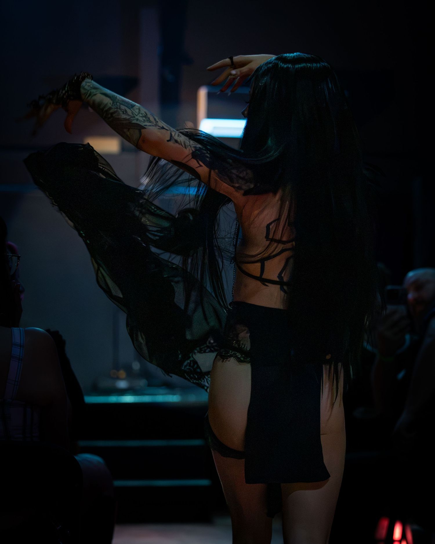 Tattooed woman in black lingerie dancing in dimly lit nightclub setting.