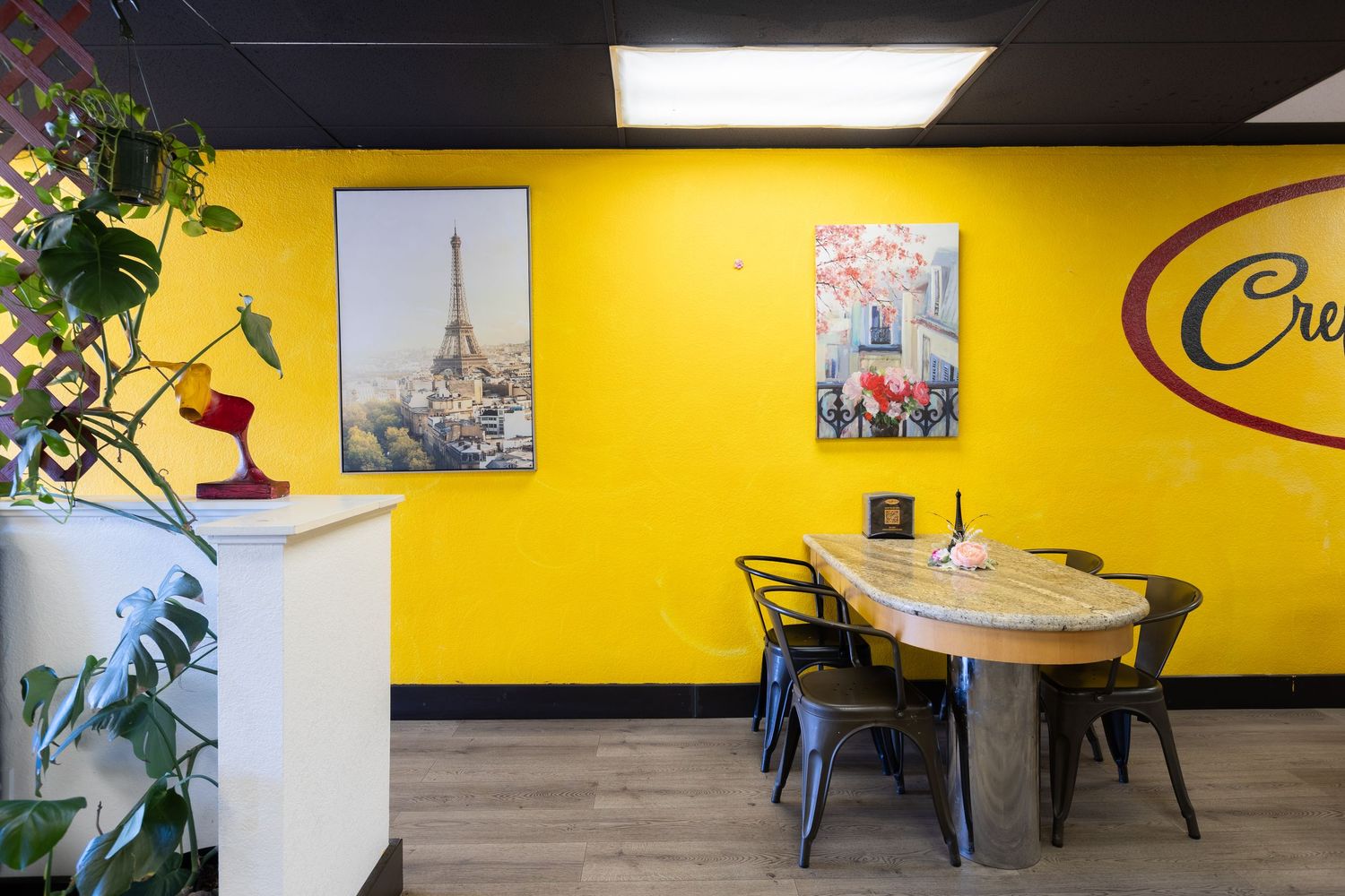 Bright yellow cafe wall with Eiffel Tower art prints, wooden dining table, and dark metal chairs in a cozy French-themed restaurant.