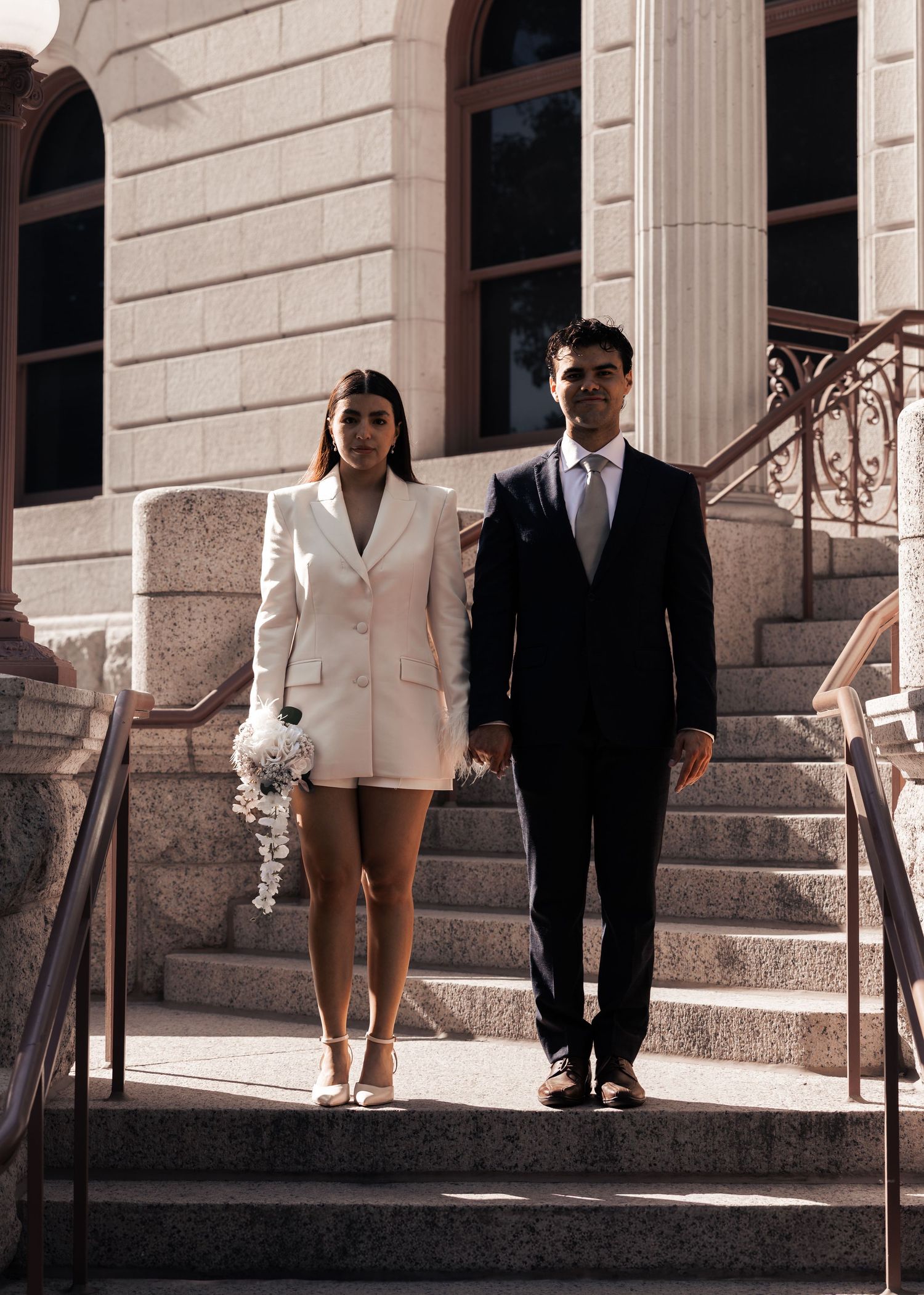 Couple in wedding attire descending stone steps, bride in white blazer dress holding bouquet, groom in black suit.