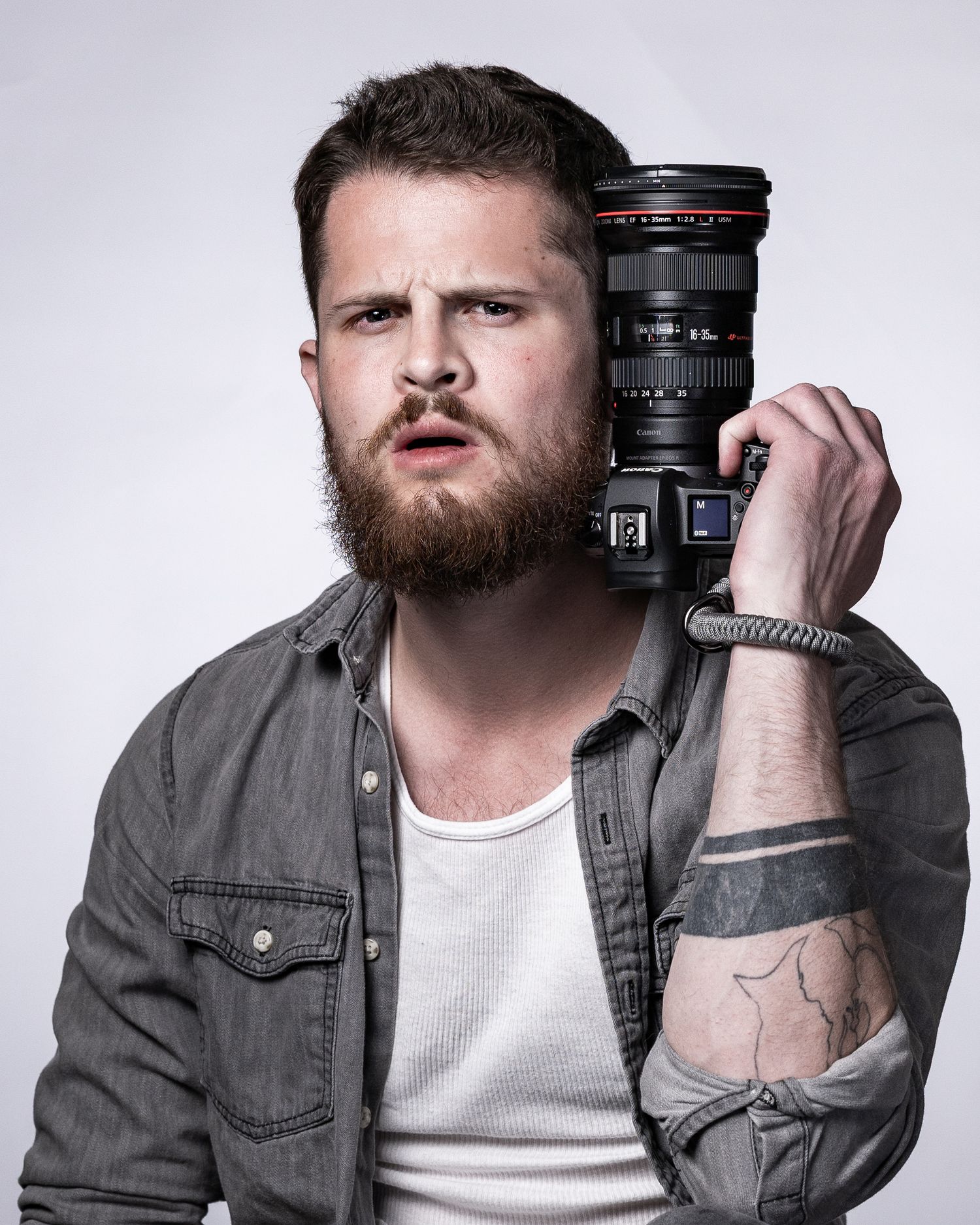 Bearded man holding a Canon DSLR camera on his shoulder, wearing a denim shirt with arm tattoos visible.