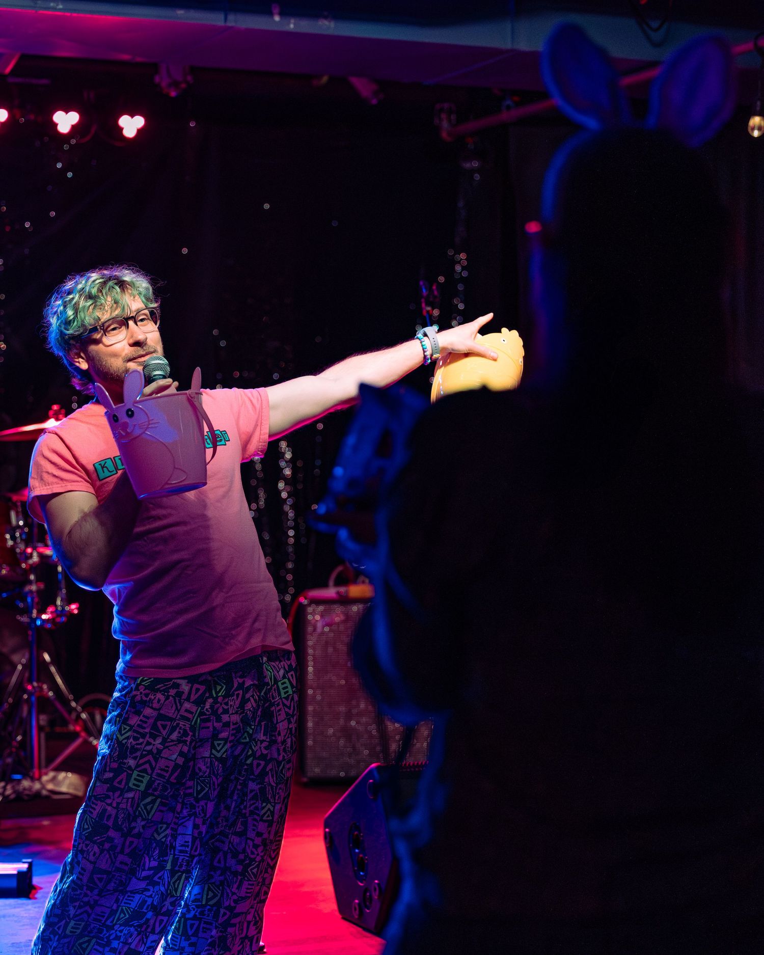 Performer in pink shirt and colorful pants gesturing toward audience member on stage.