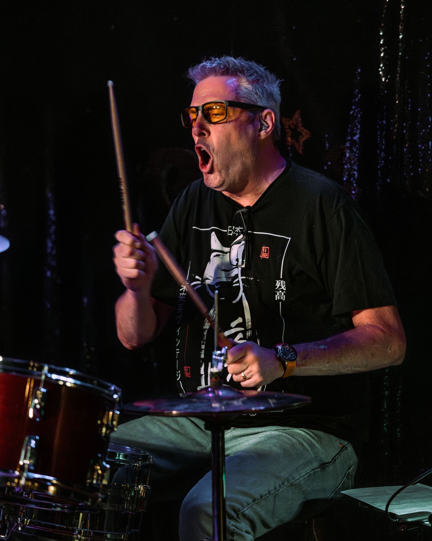 Energetic drummer with orange glasses plays drums with open mouth expression under stage lighting.