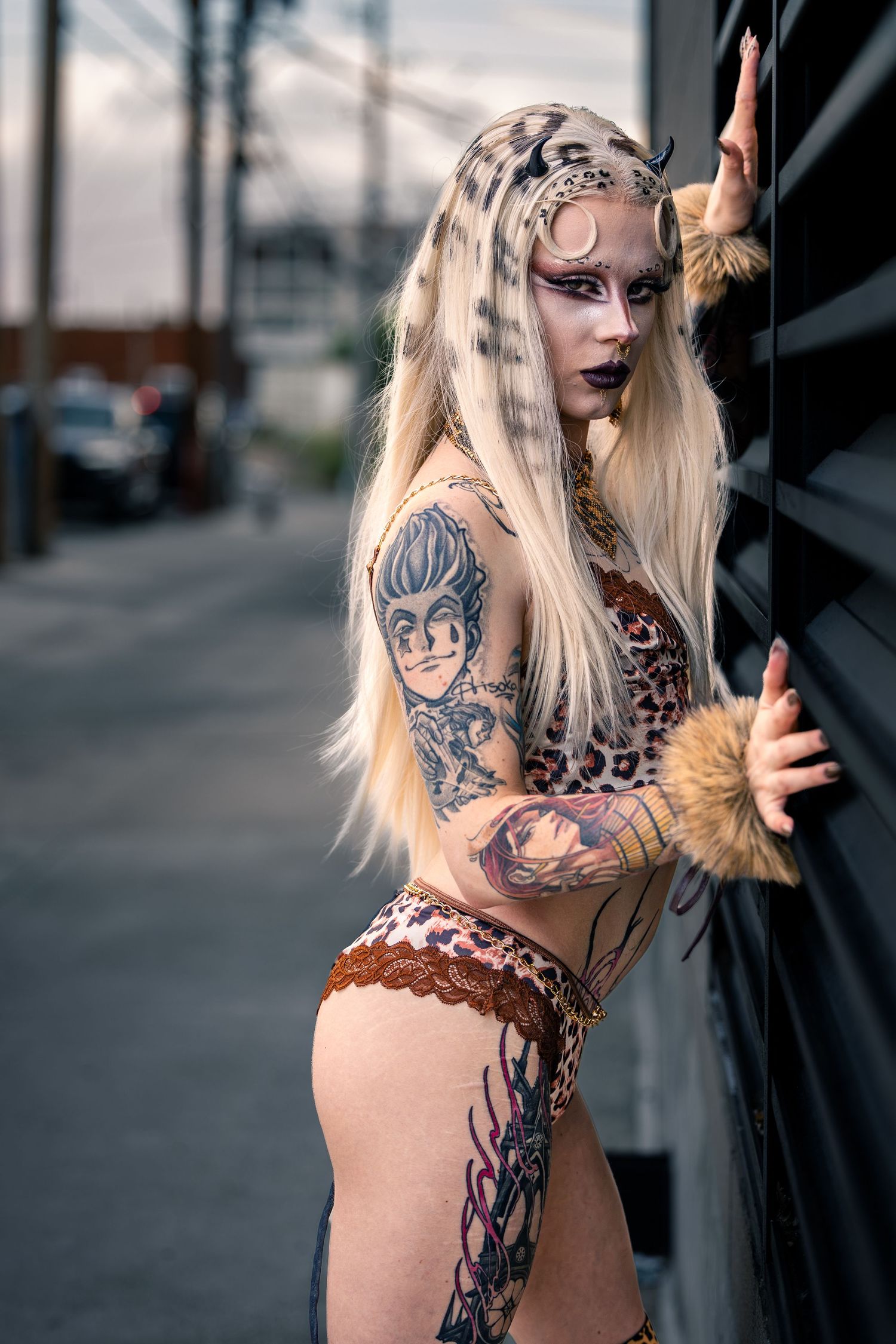 Tattooed blonde woman in leopard costume with horns leaning against wall.