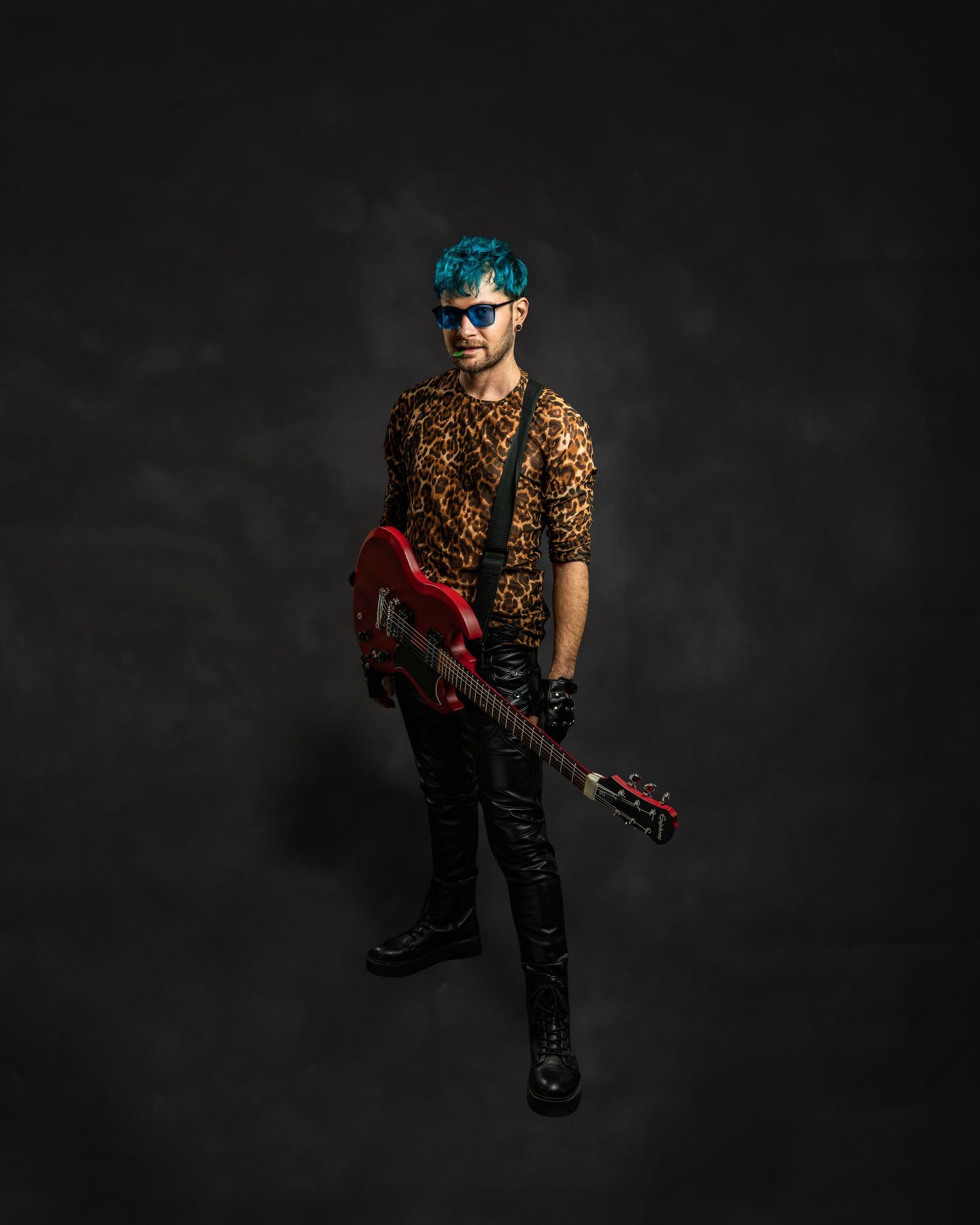 Teal-haired guitarist in leopard print shirt holds red electric guitar wearing sunglasses in dark studio.