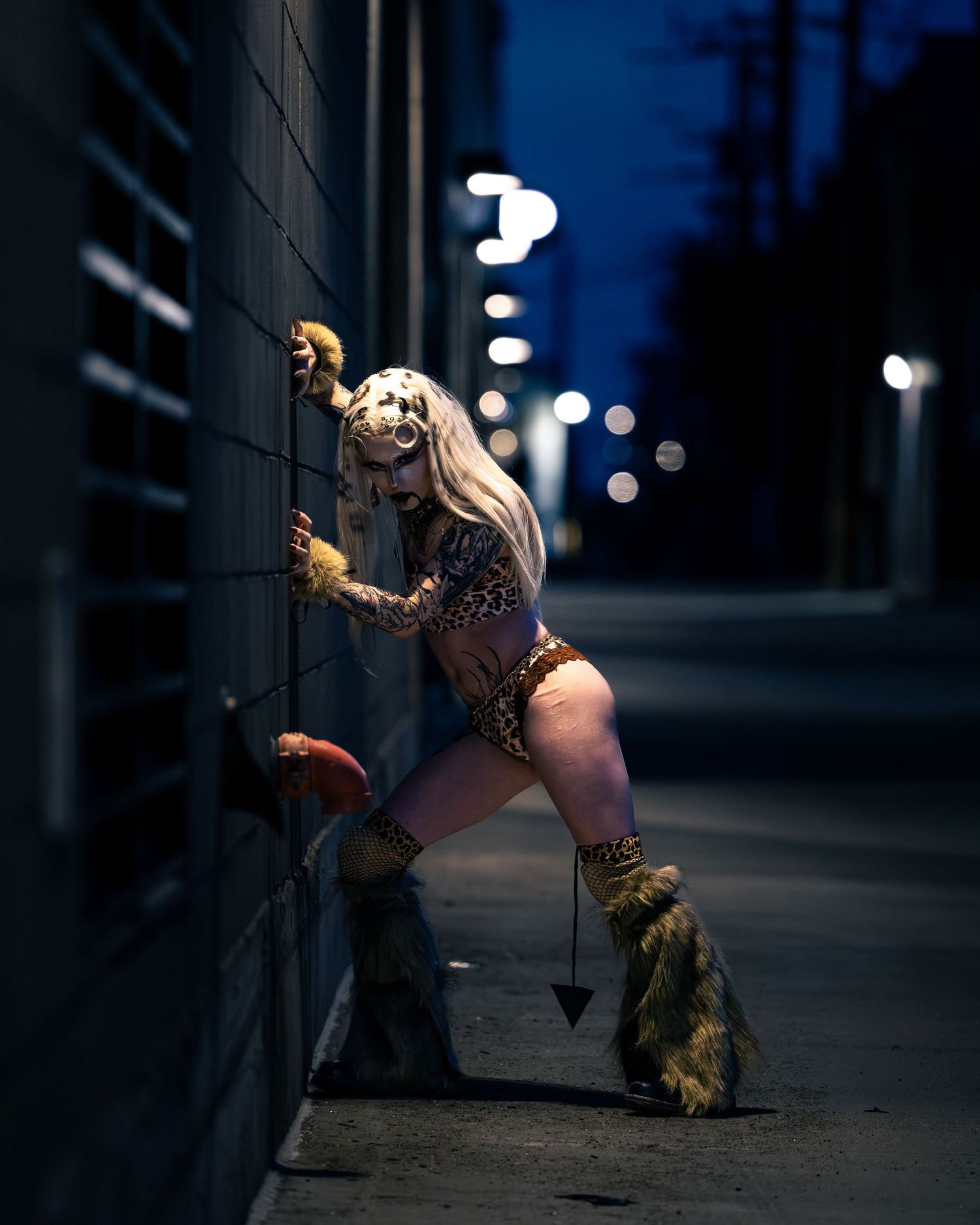 Tattooed blonde in leopard print outfit with fur boots leaning on wall at night.