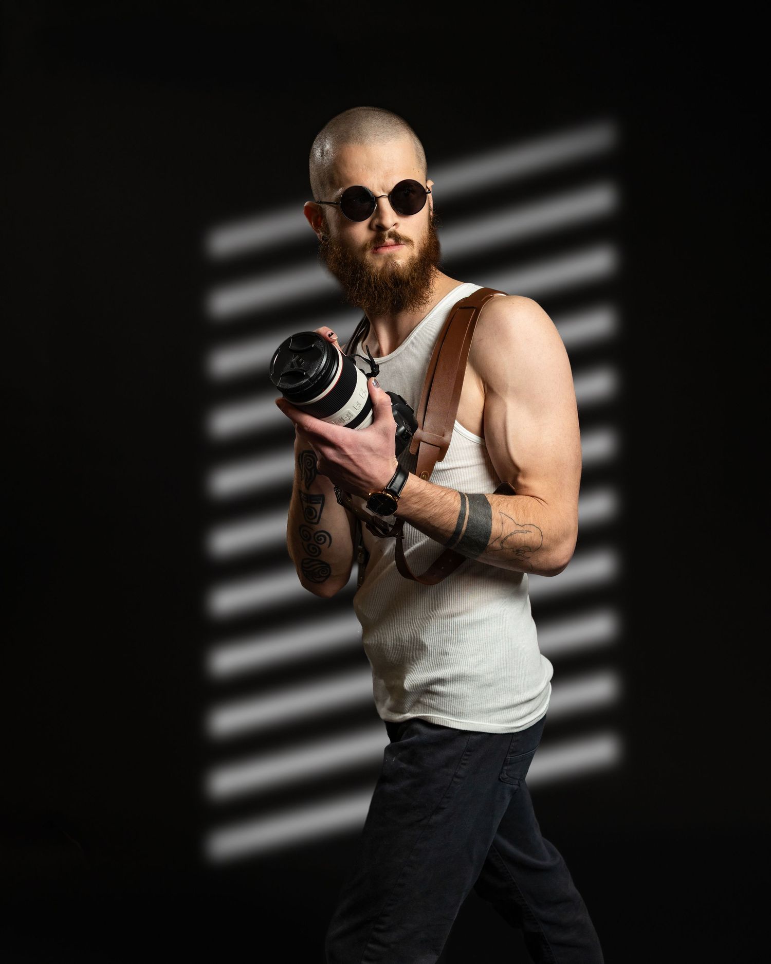 Professional photographer in a white tank top and sunglasses works with camera equipment against dark background.
