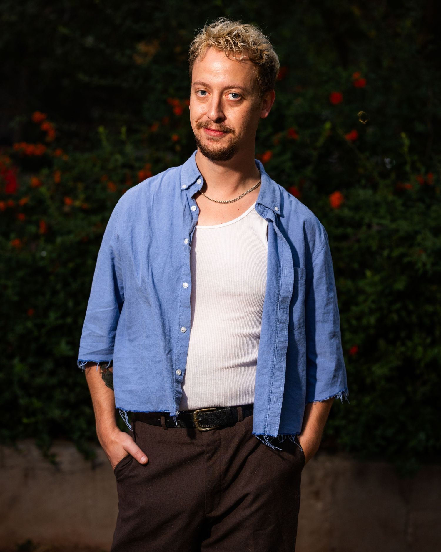 Person in blue button-down shirt and dark pants stands outdoors against dark background with red flowers.