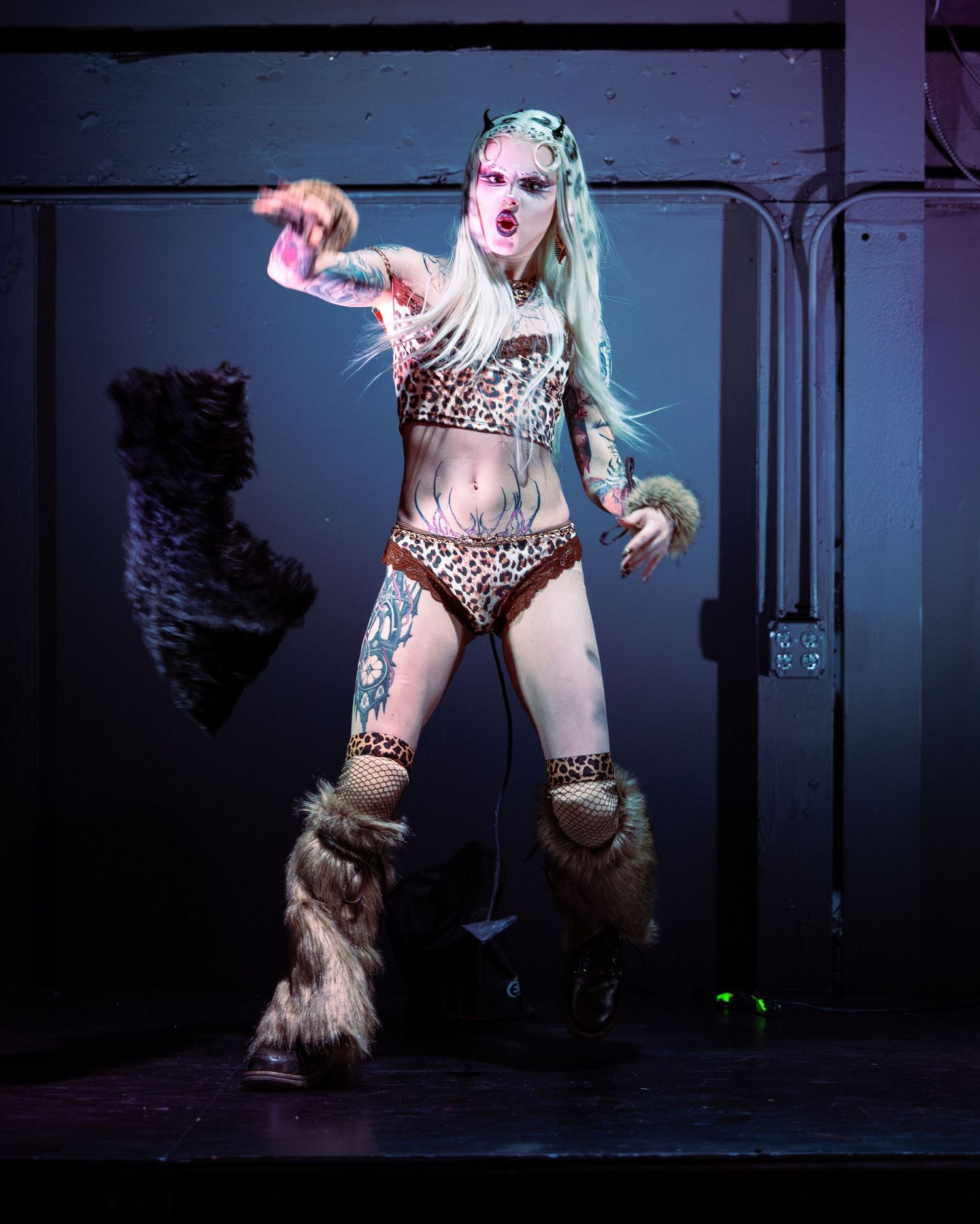 Tattooed performer in leopard print costume with fur boots and devil horns poses on stage under blue lighting.