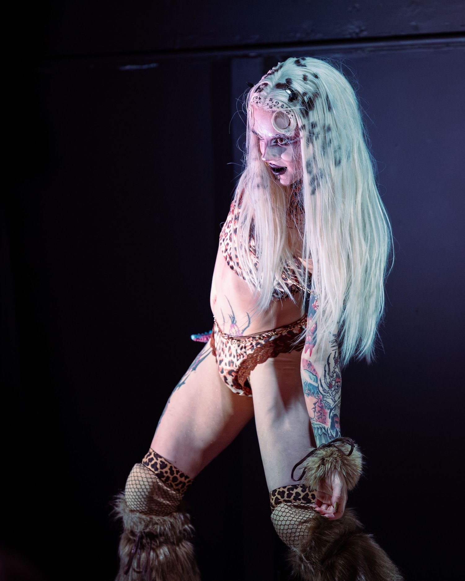 Tattooed burlesque performer in leopard print outfit with dalmatian face paint and fur boots on stage.