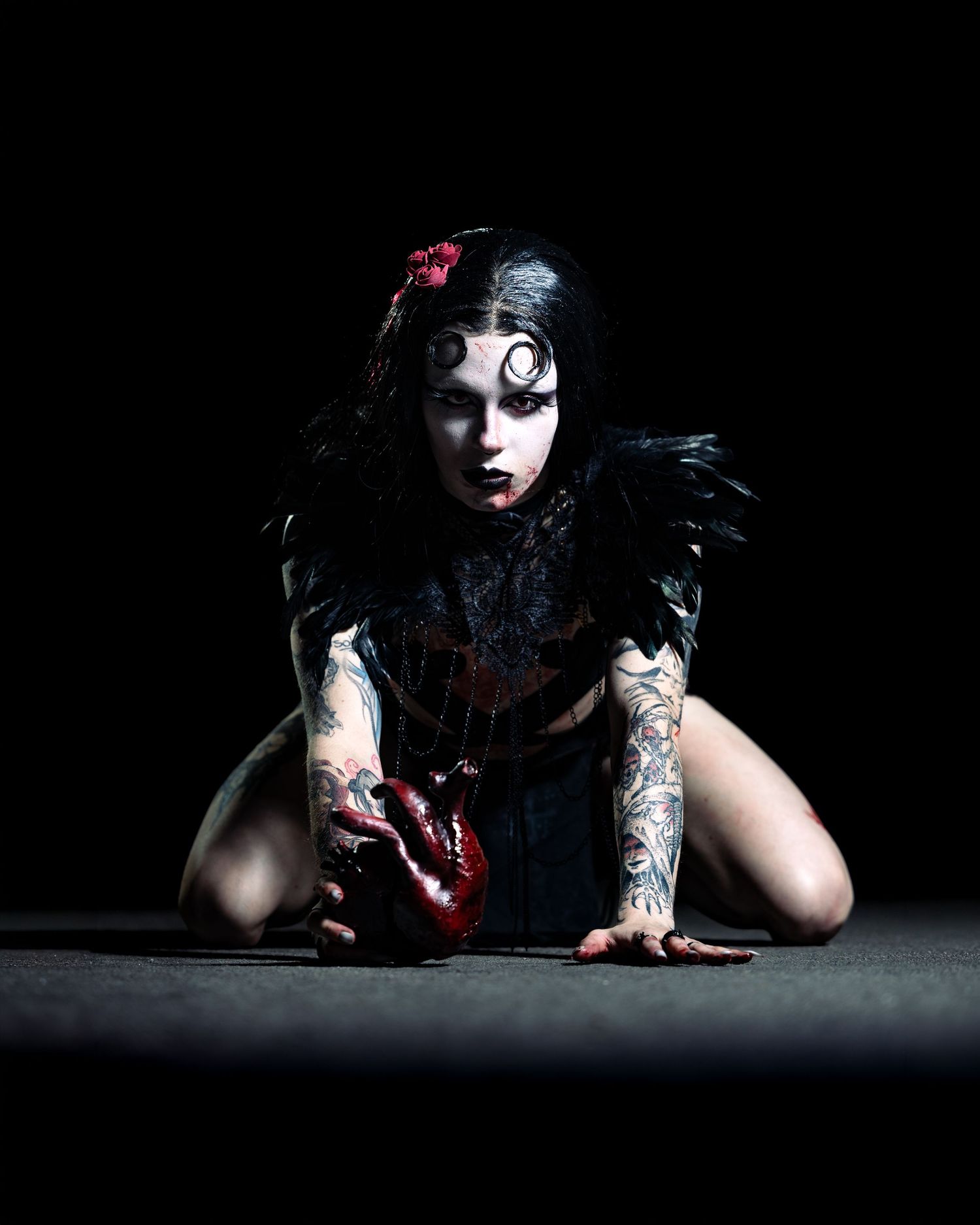 Gothic woman with tattoos, dark makeup, black feathers, and dramatic horror-themed styling poses on a dark stage.
