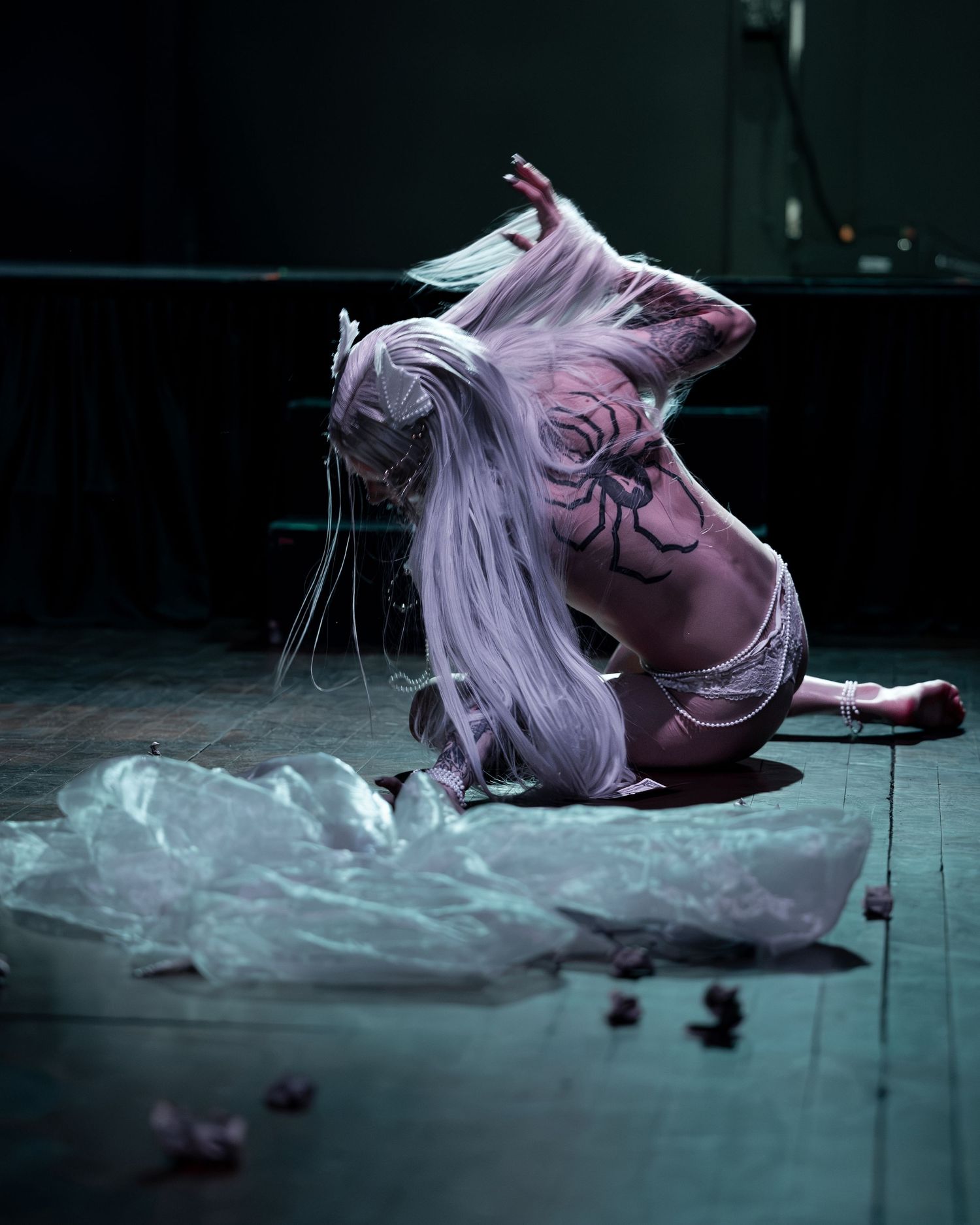 Performer with long white hair and body paint kneels dramatically on dark stage floor surrounded by sheer fabric.