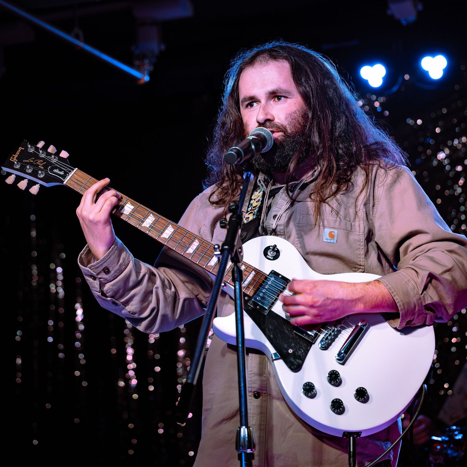 Bearded guitarist with long hair playing a white Les Paul guitar at a microphone.