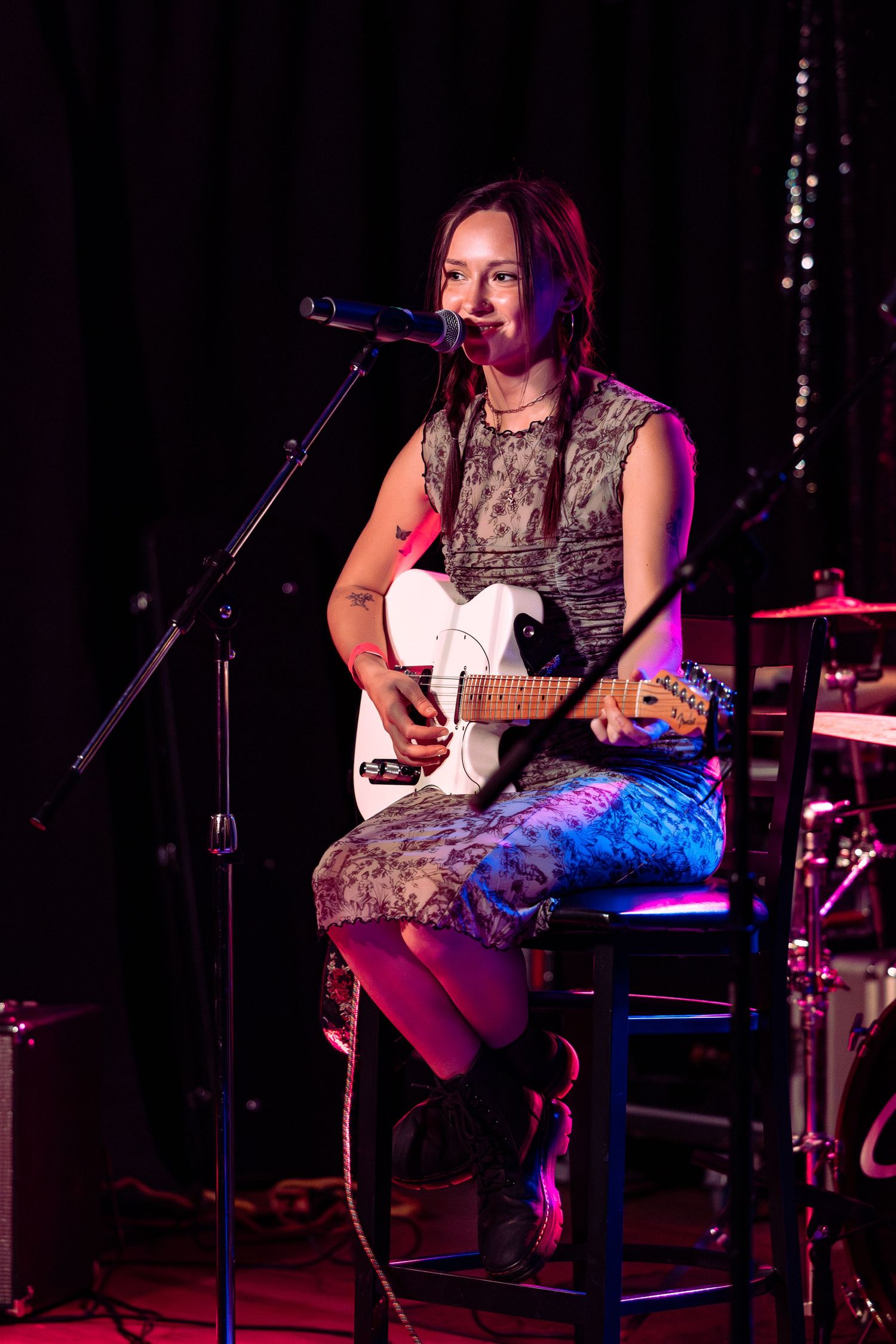 Woman playing white electric guitar while singing into microphone on stage under pink stage lighting.