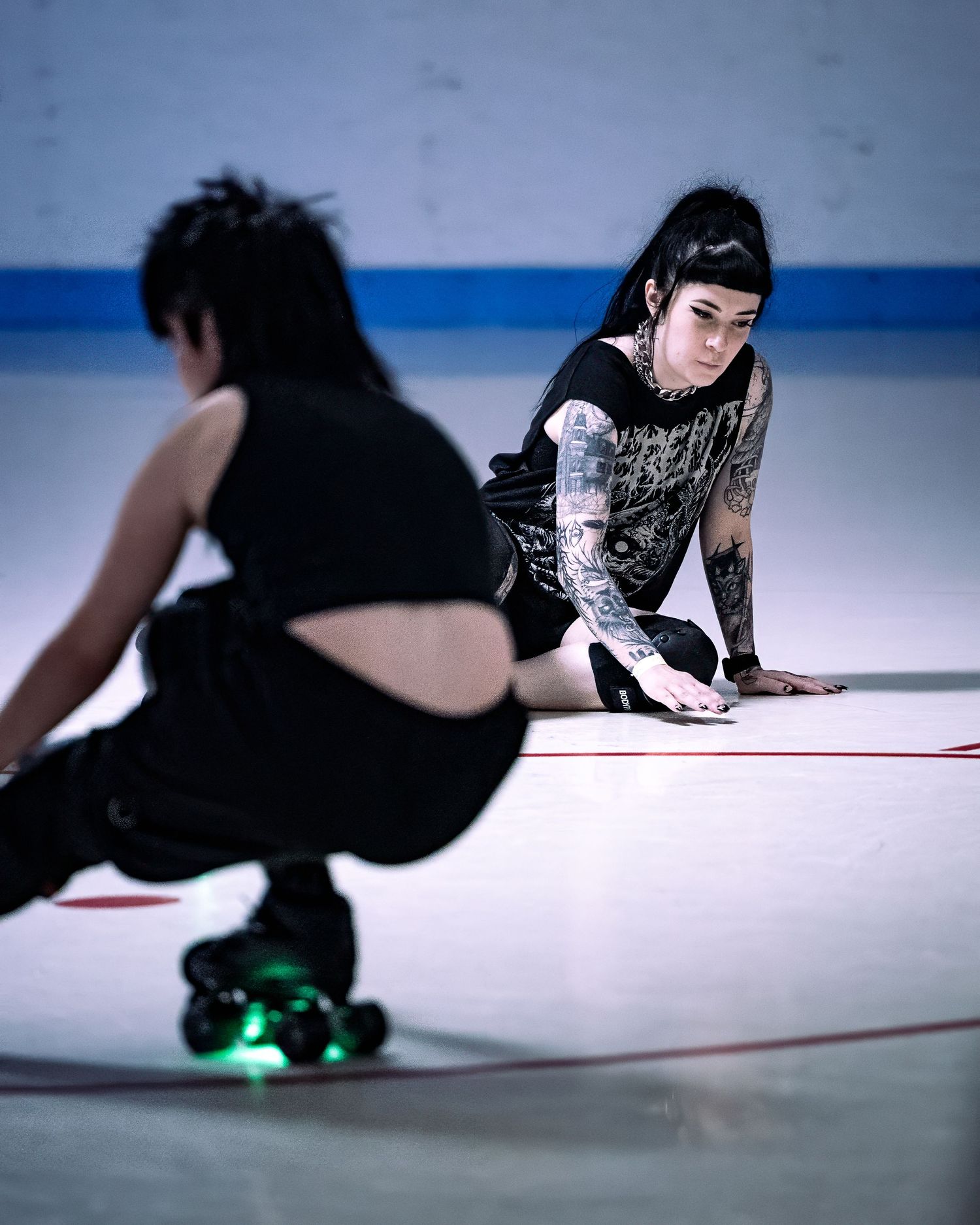 Tattooed roller derby skater crouches on rink floor in black outfit with green-lit skates during practice.