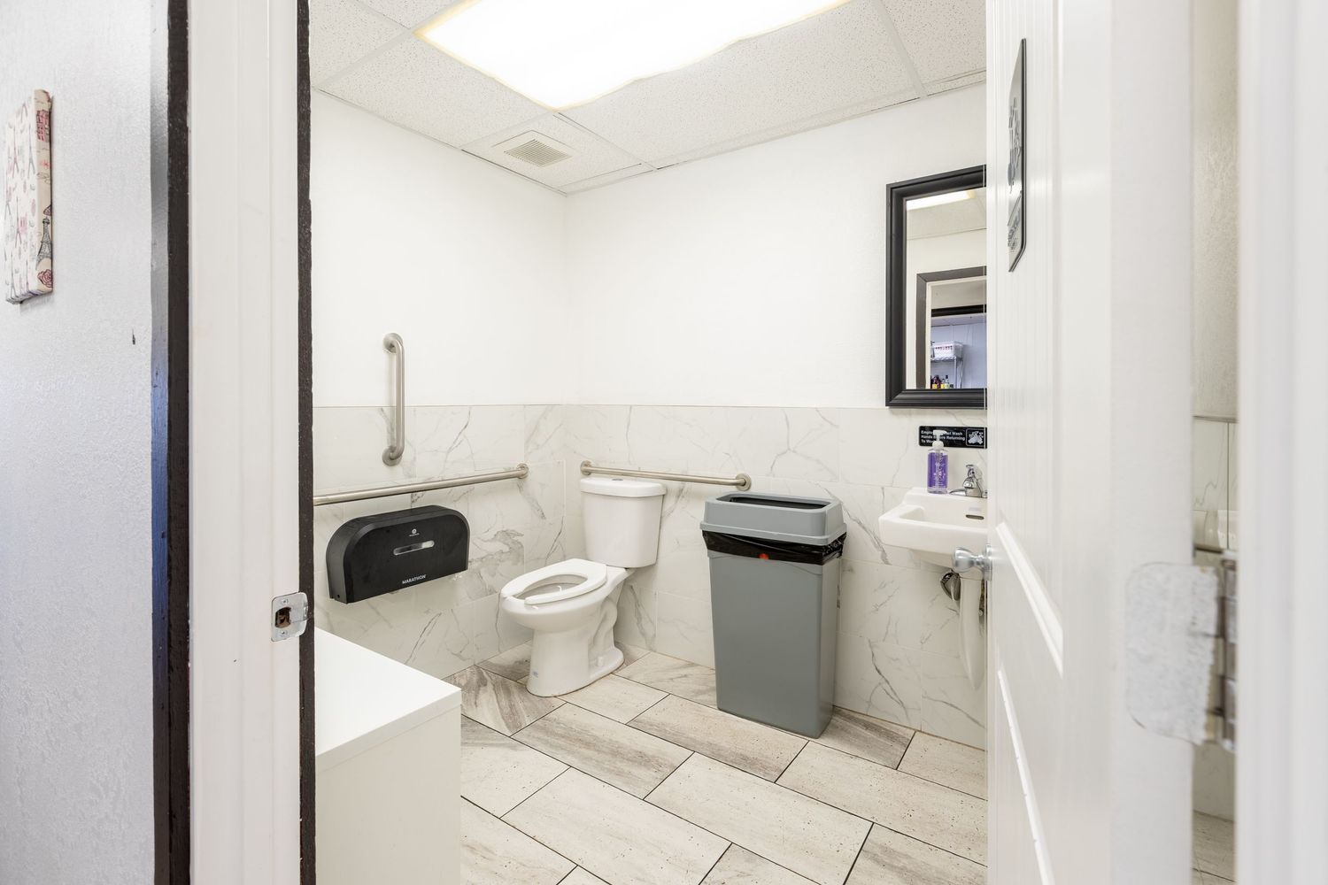 Accessible commercial restroom with marble tile walls, grab bars, toilet, trash can, and sink with mirror.